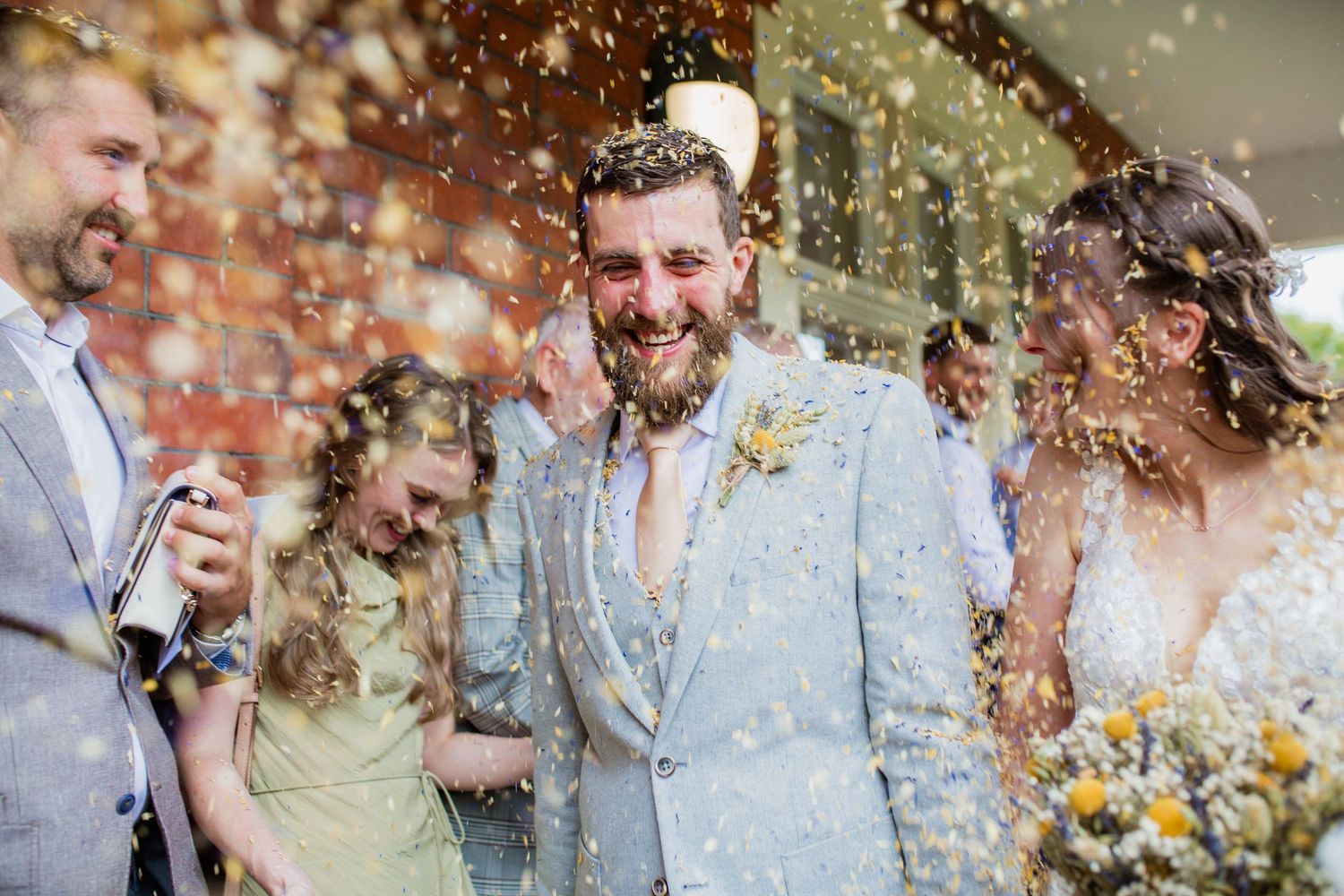 Happy wedding celebration with guests throwing confetti in natural outdoor setting.