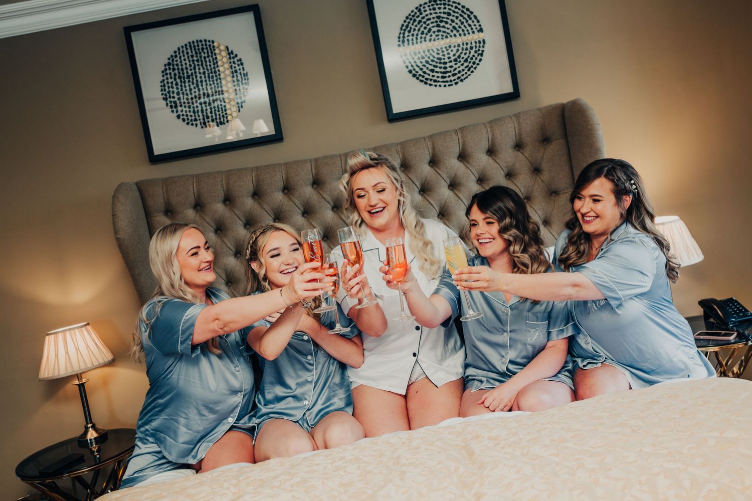 Group in matching blue robes toasting with champagne glasses on a luxurious bed.