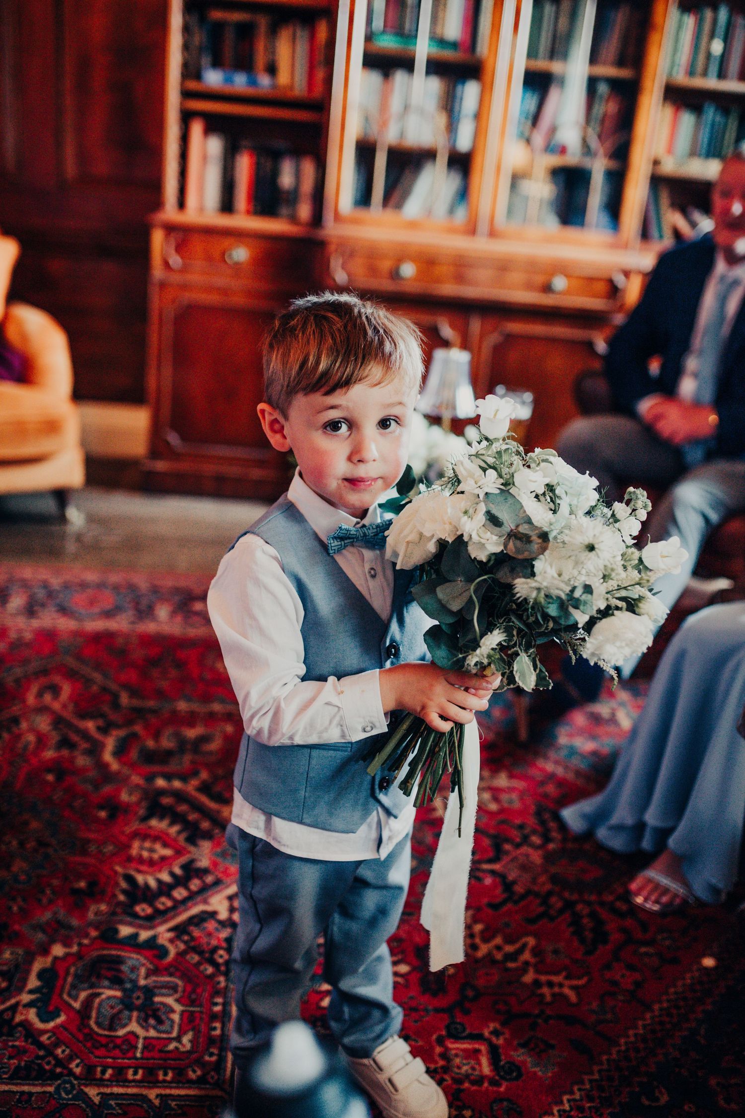 Young ring bearer in a gray vest holds a large white floral bouquet during an elegant indoor wedding ceremony.