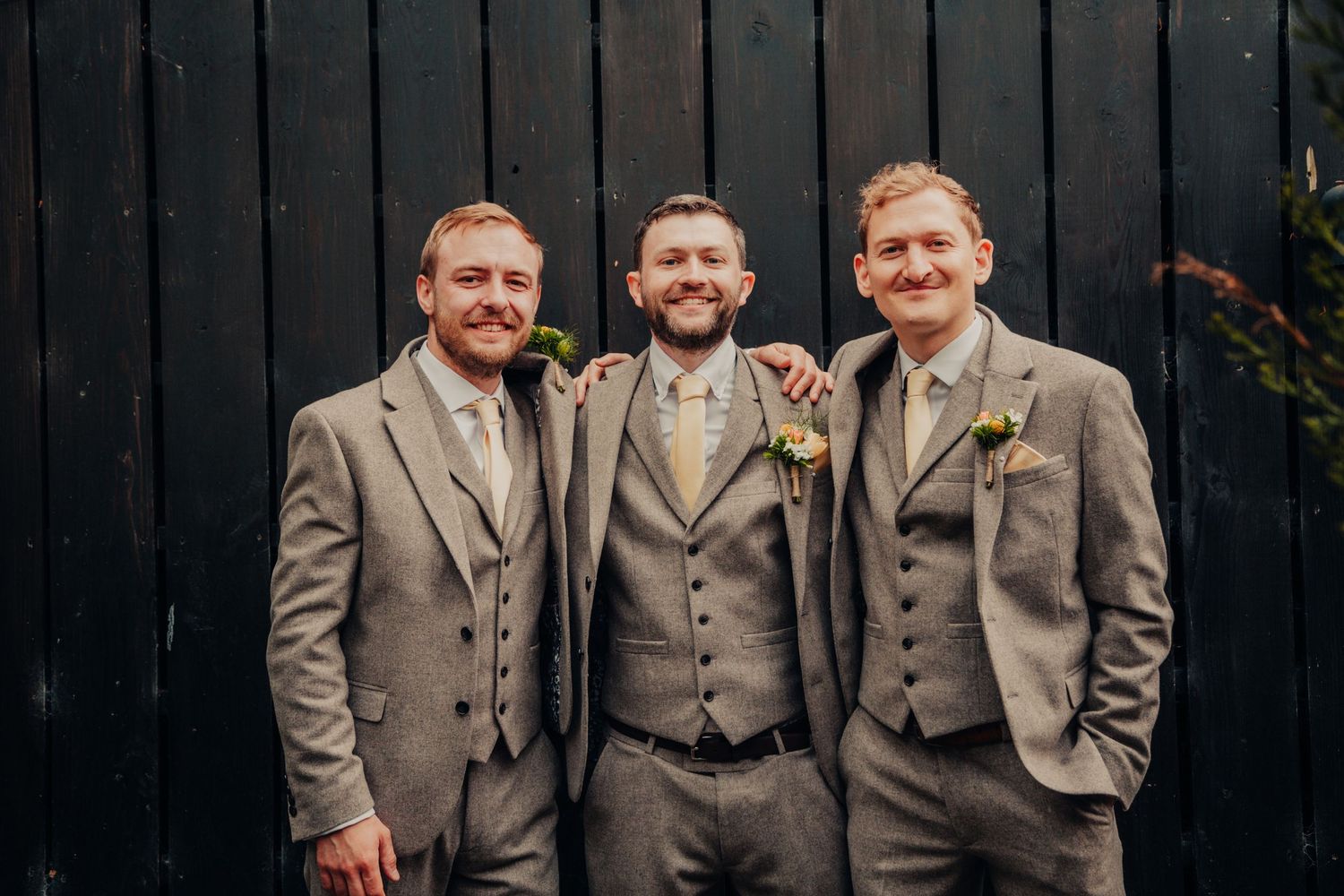 Three groomsmen in matching gray suits pose together smiling against a dark wooden wall backdrop.