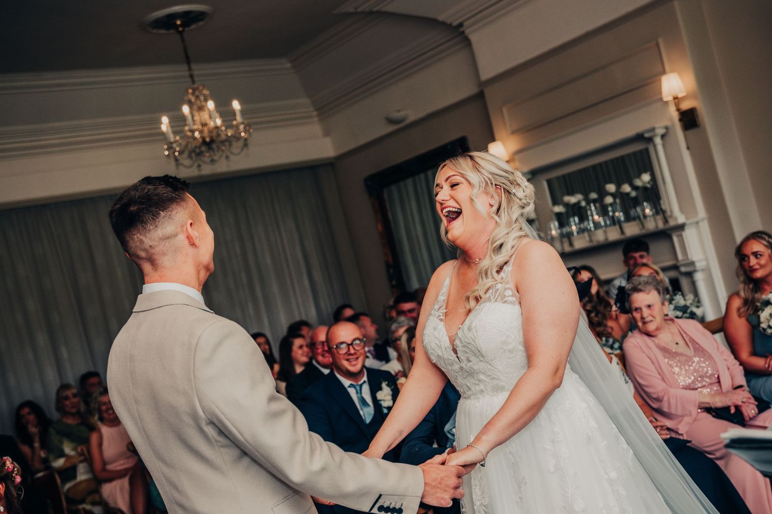Couple laughing joyfully during their wedding ceremony in a chandelier-lit venue.