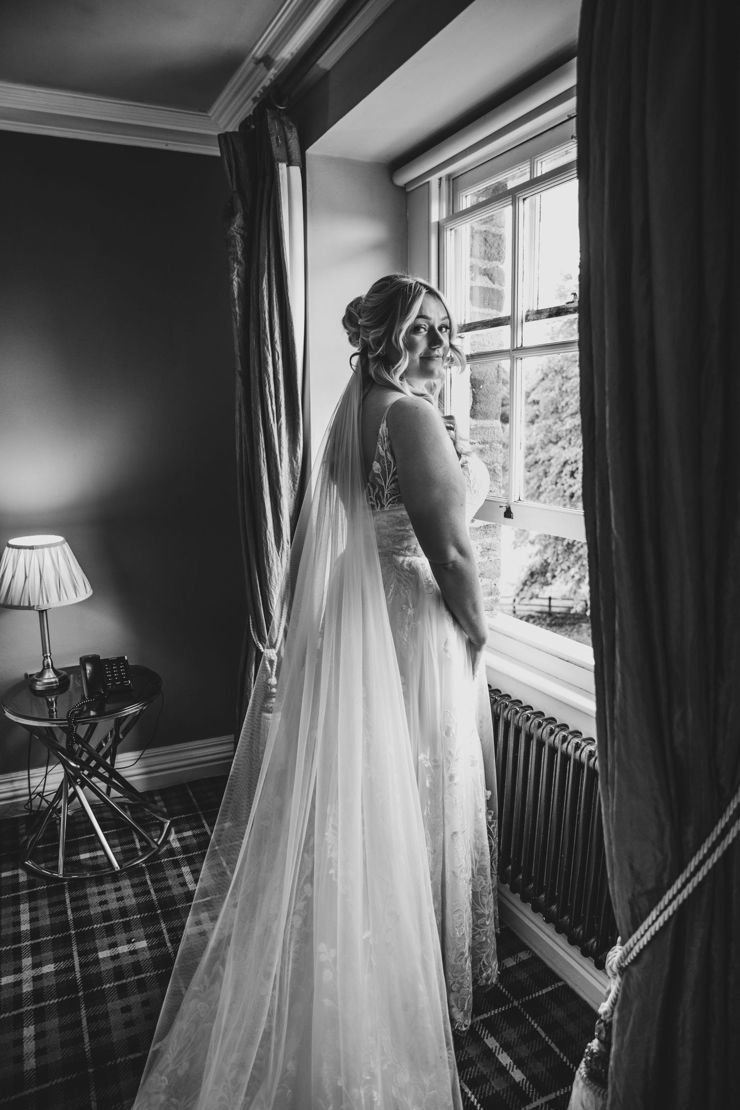 A bride in a flowing white gown and veil gazes out a window in a classic black and white portrait.