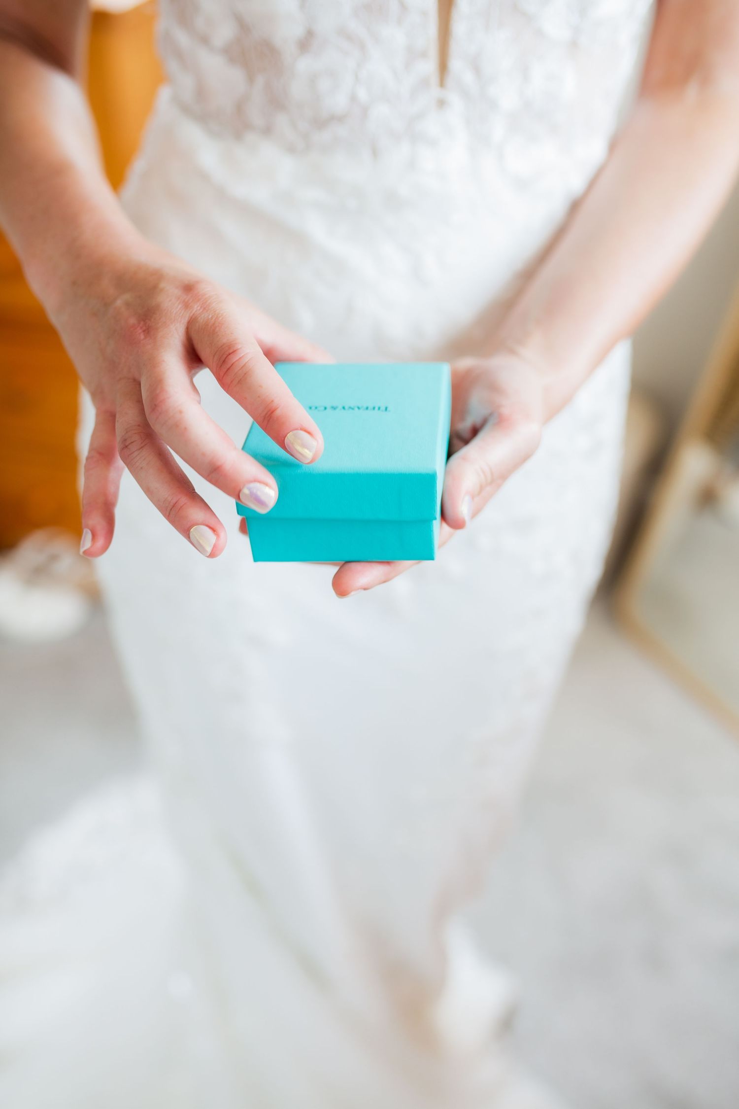 Close-up view of hands holding a small turquoise blue gift box against a white wedding dress background.
