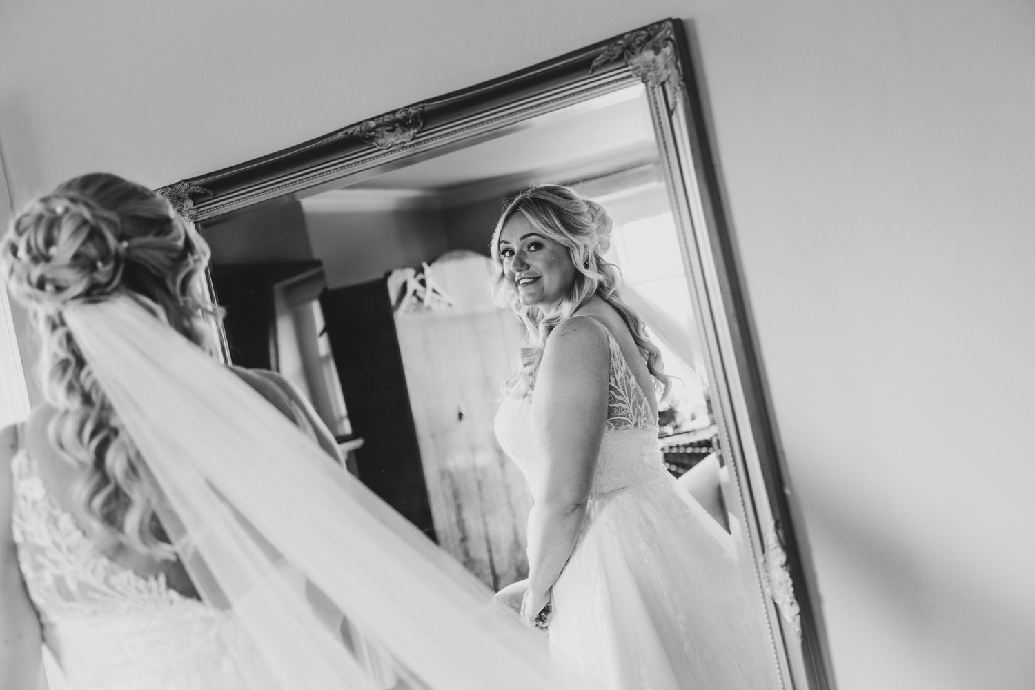A bride admires her reflection in an ornate mirror while wearing a flowing white wedding dress.