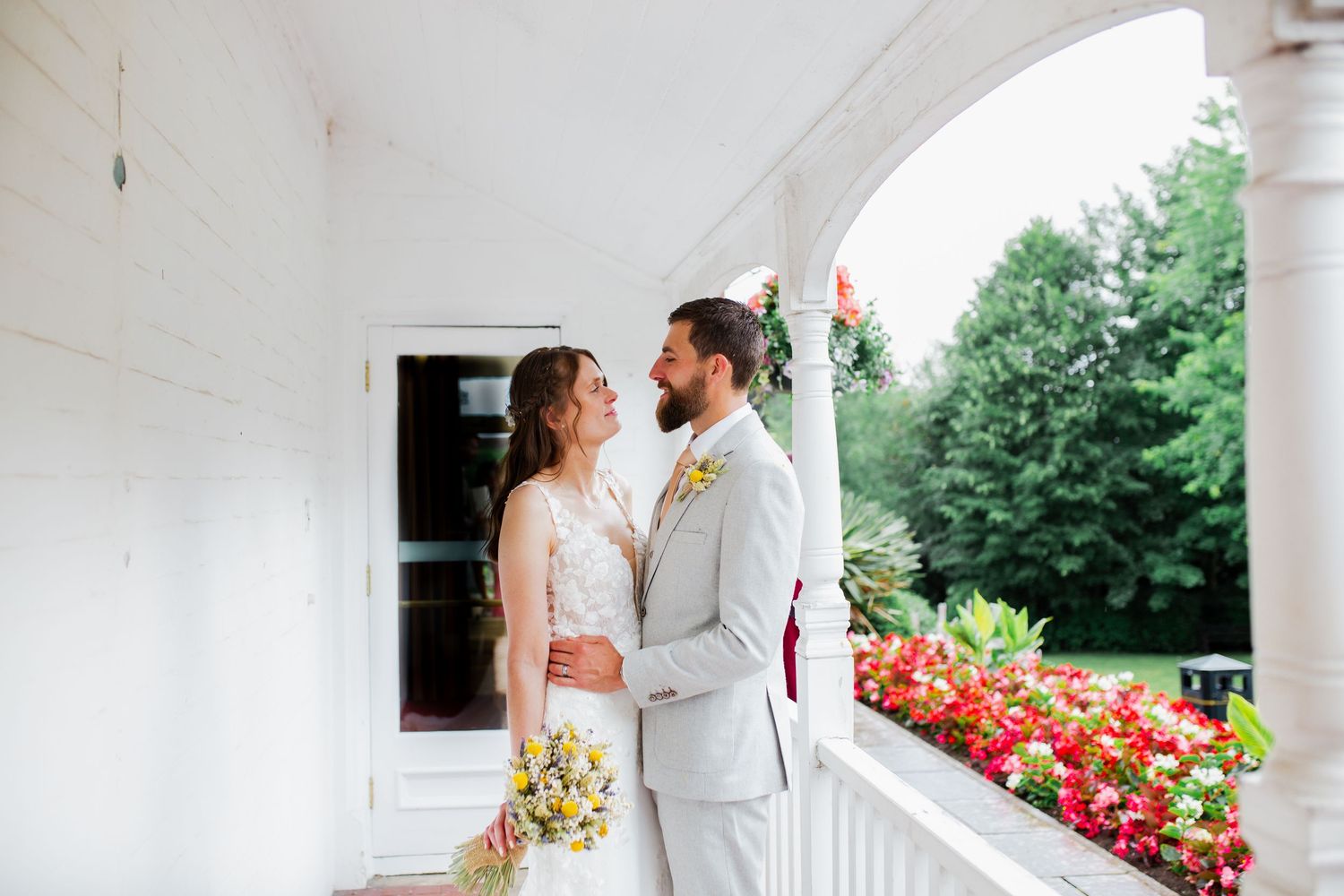 Romantic moment between newlyweds on a white porch with red flowers in the background.