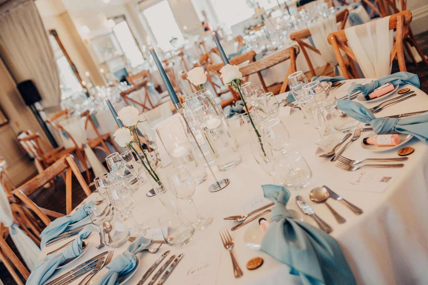 Rustic wedding reception tables set with light blue linens, wooden chairs, floral arrangements and soft ambient lighting.