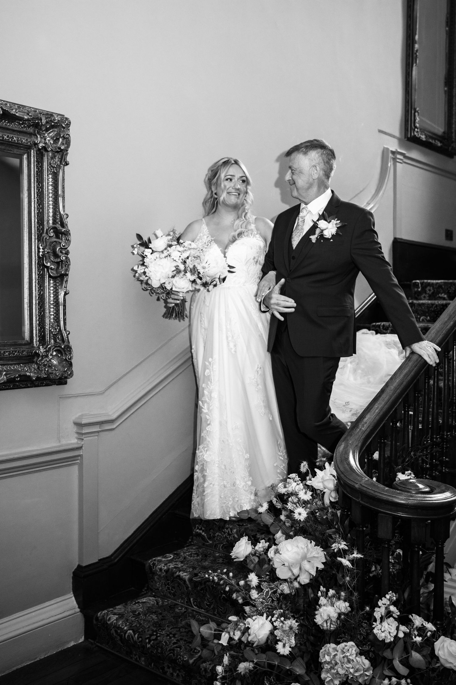 A couple in wedding attire poses on an elegant staircase decorated with flowers and a vintage framed mirror.