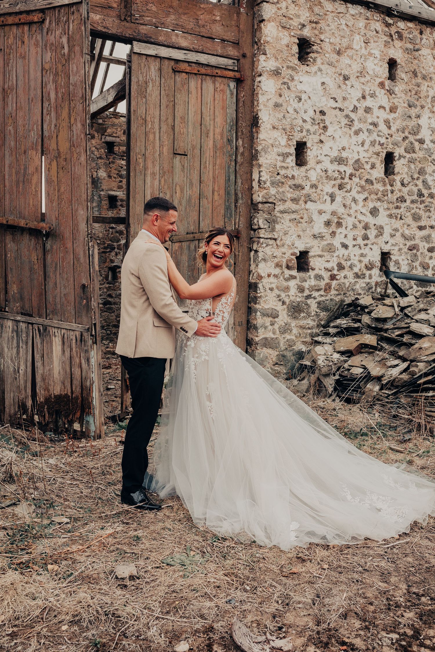 A couple in wedding attire poses in front of rustic wooden doors and stone wall at a barn or farmhouse setting.