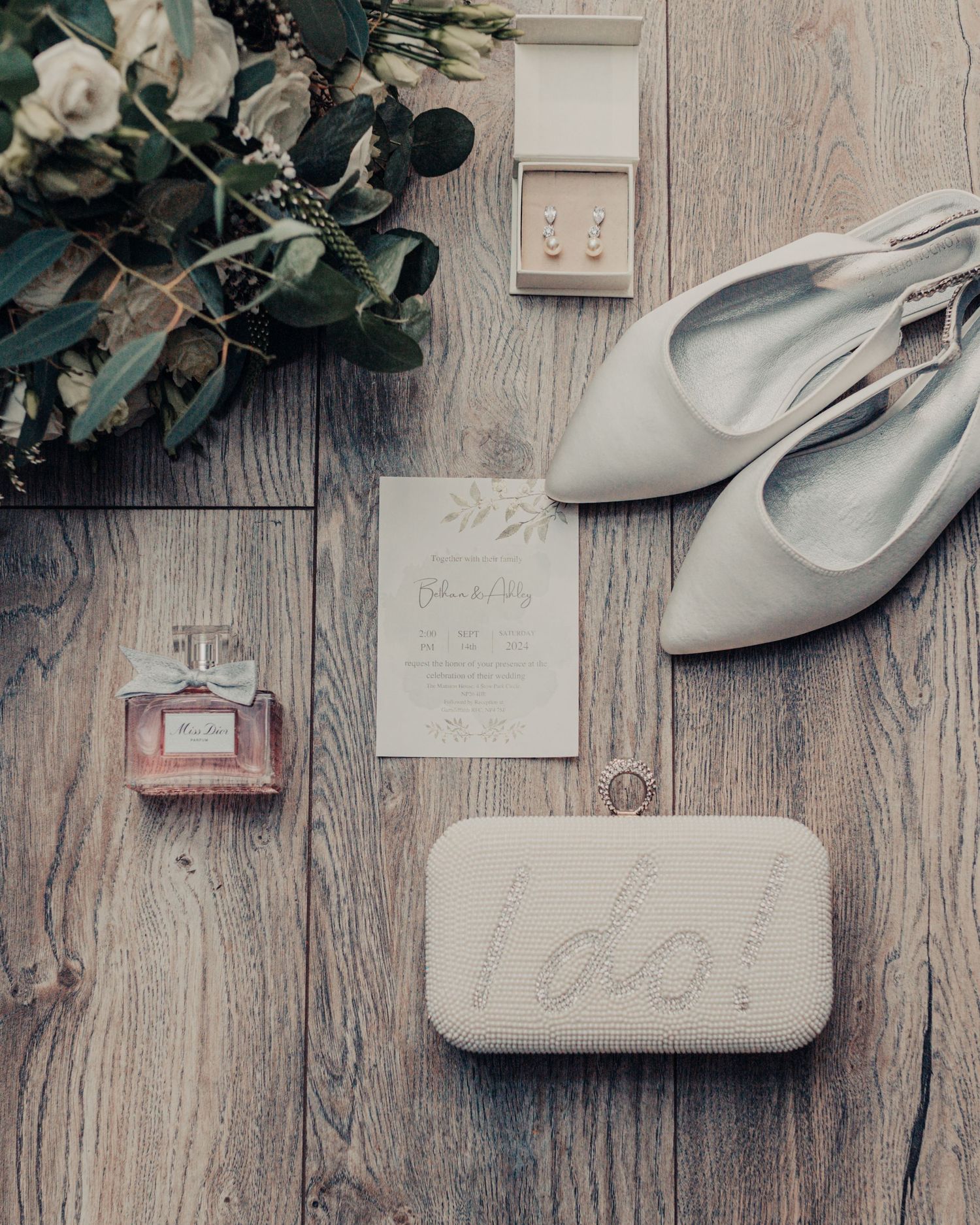 Wedding details flatlay with silver shoes, perfume bottle, and floral arrangement on wooden surface.