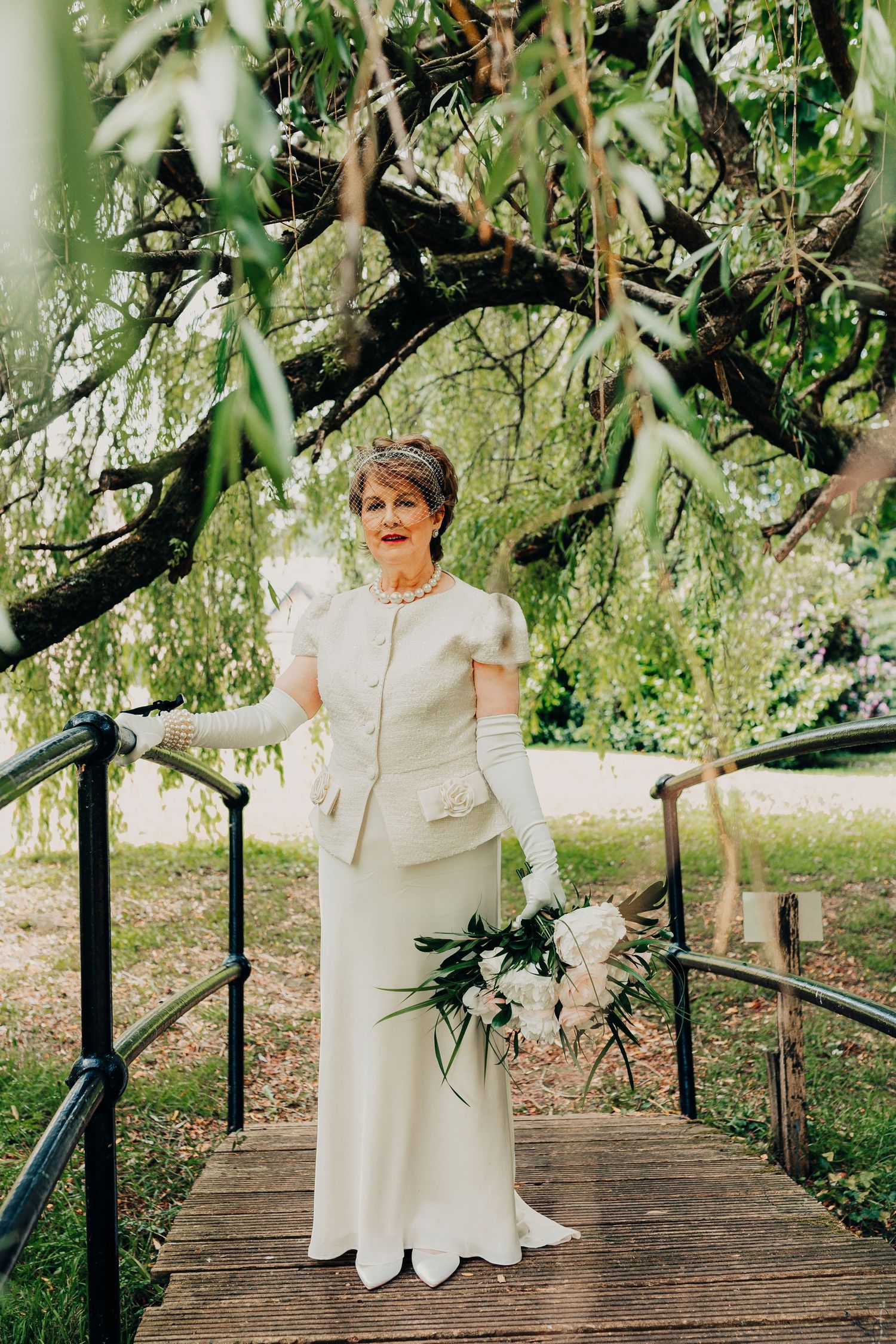 Woman in elegant cream coloured dress and gloves poses on wooden bridge surrounded by greenery.