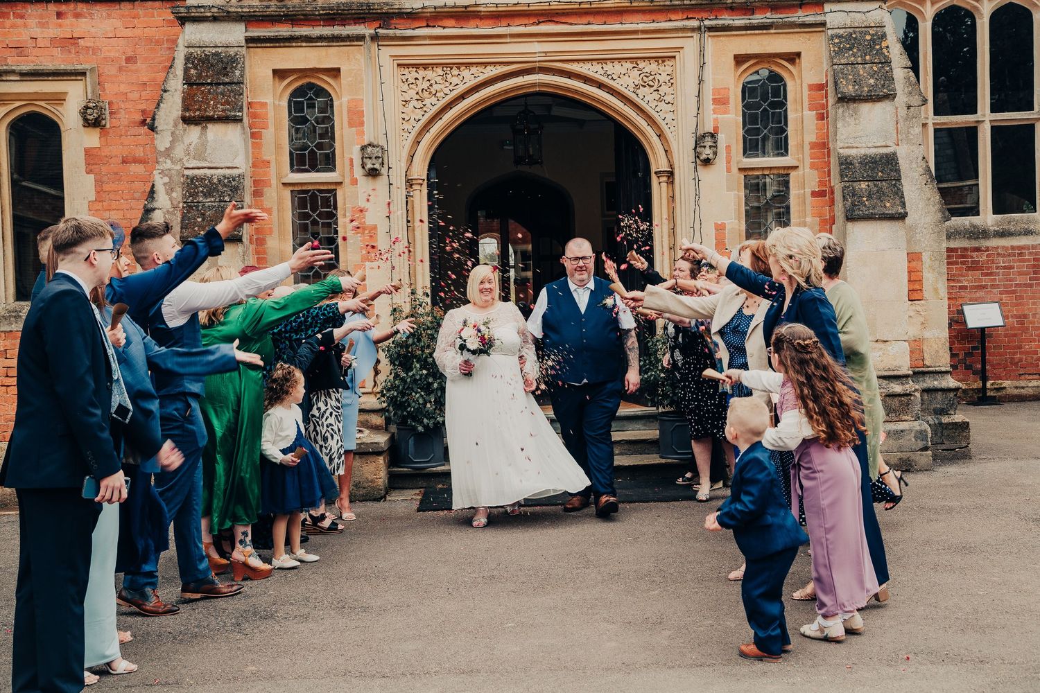 A line of guests throws confetti outside a historic brick building entrance during a wedding celebration.