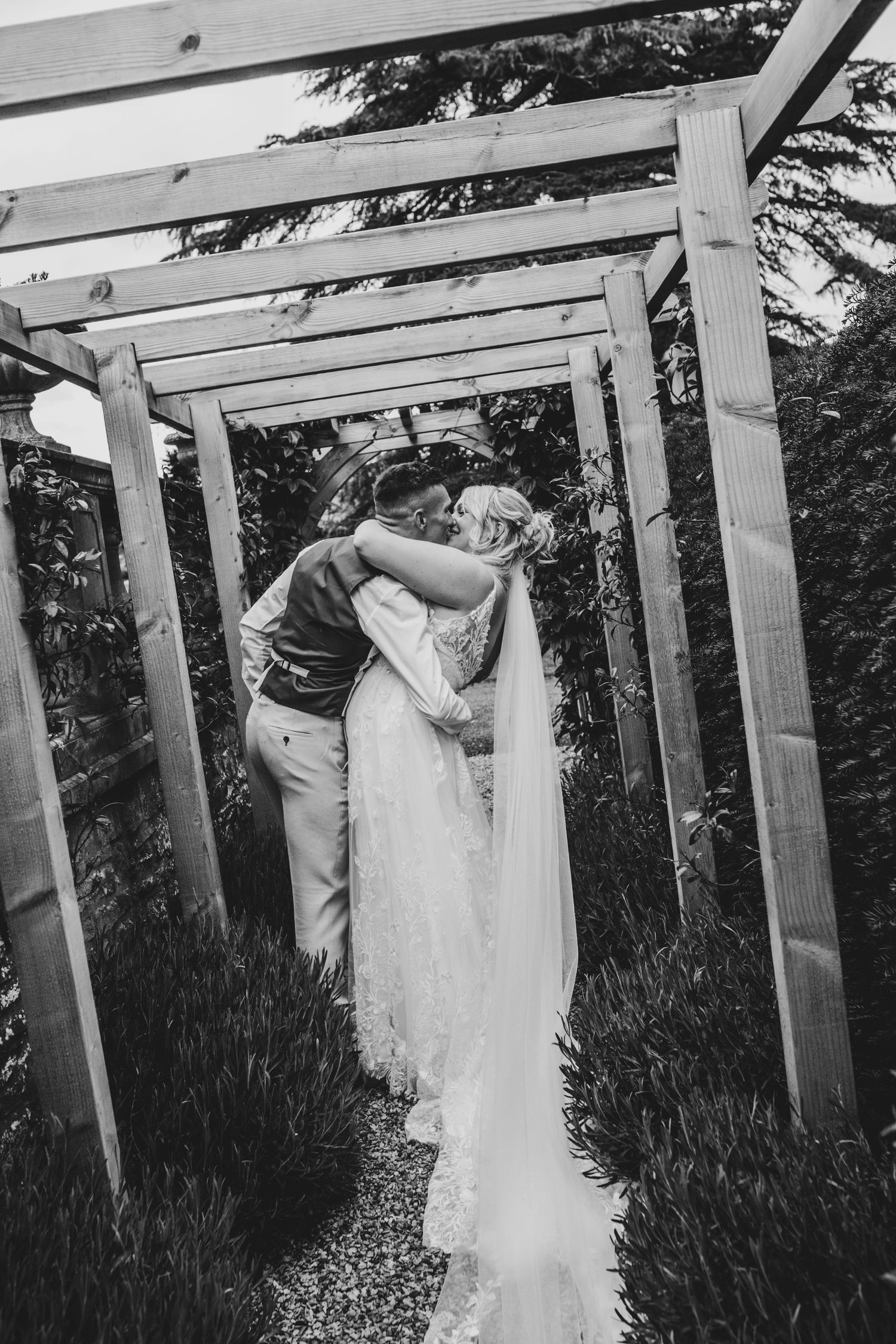 Wedding couple poses under a rustic wooden pergola surrounded by flowers.