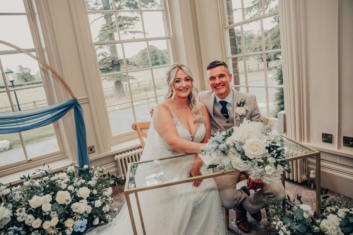 Newlyweds posing by large windows with elegant white floral arrangements.