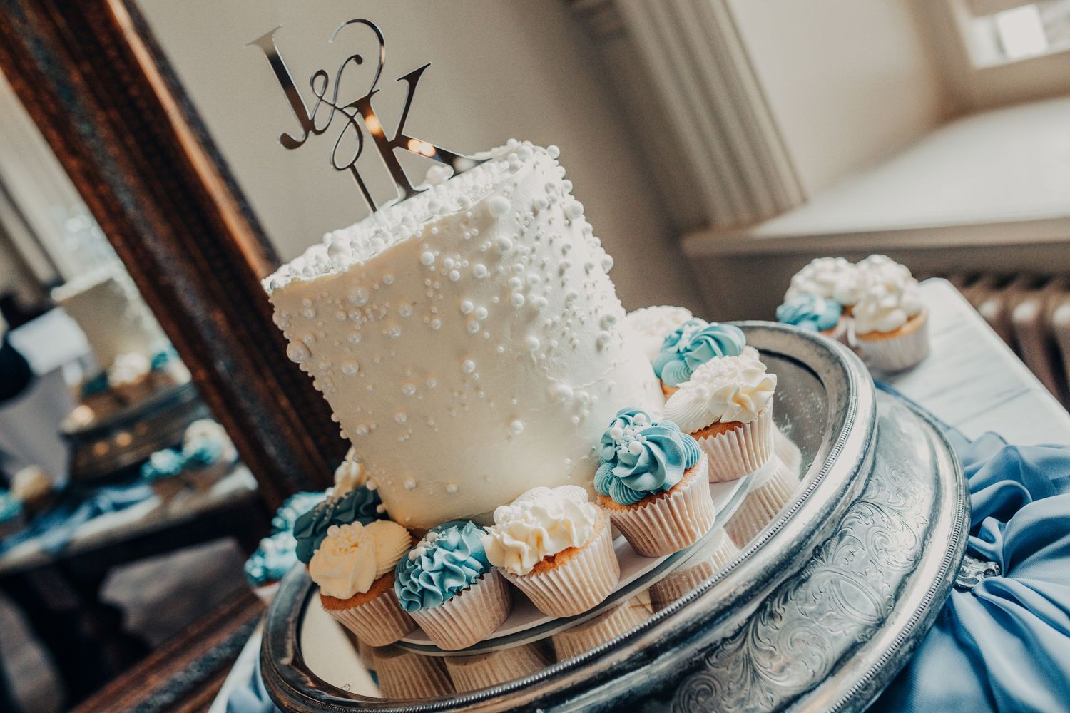 White wedding cake with blue and cream colored cupcakes arranged on a silver cake stand for reception display.