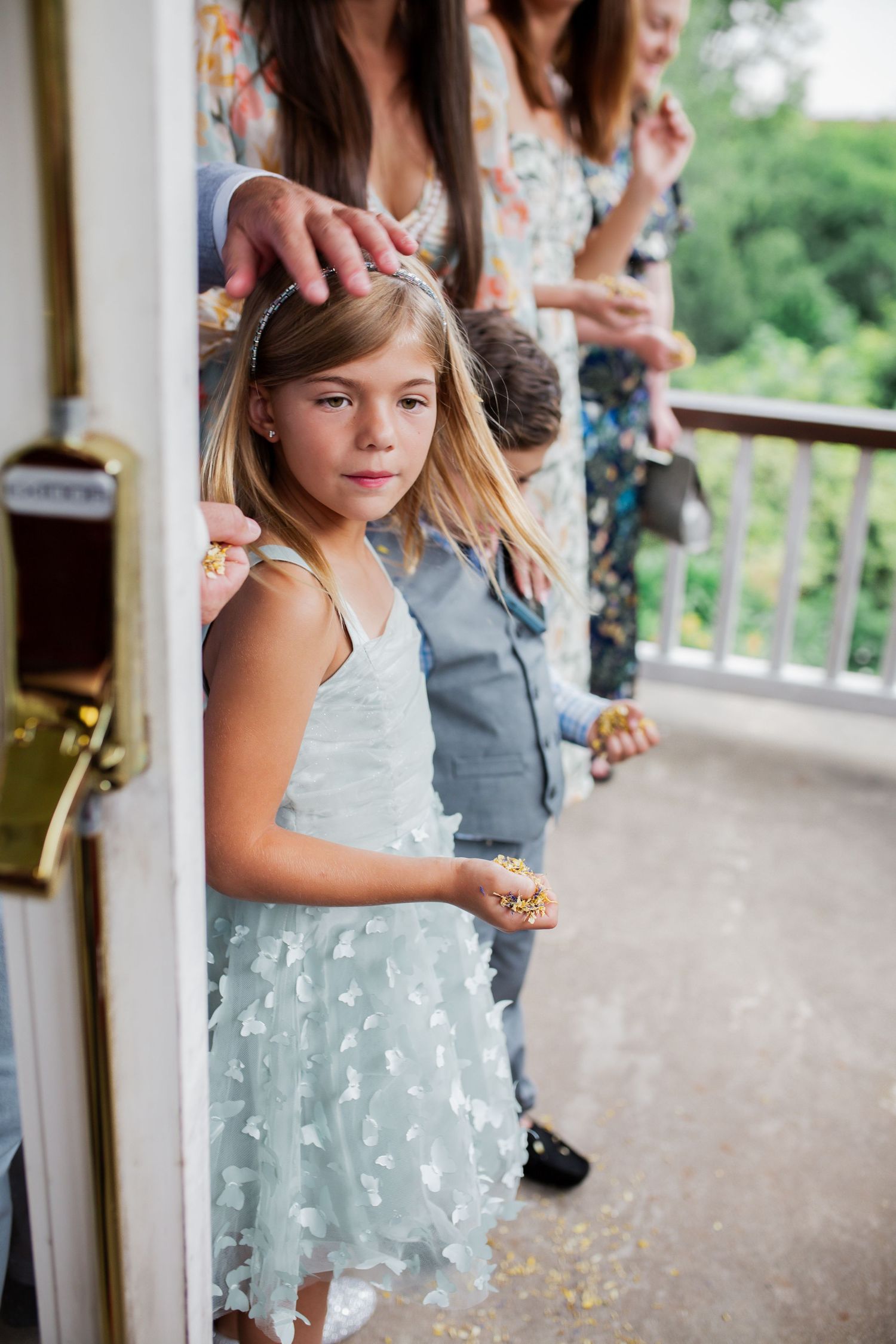Young flower girl in light blue dress with floral details on outdoor porch.