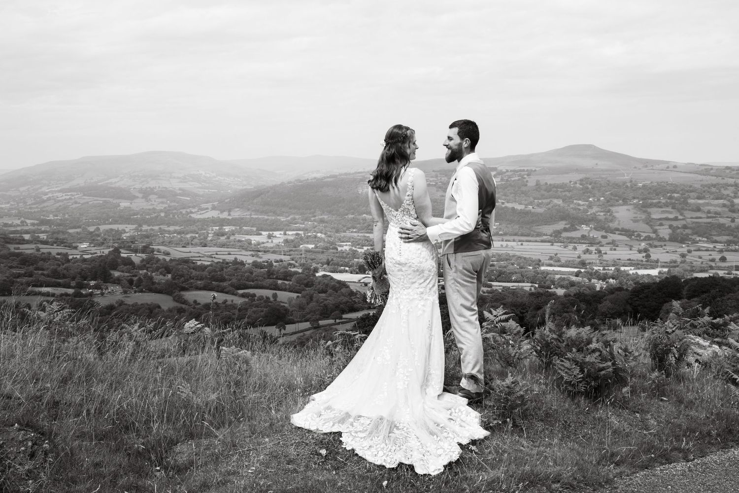 Romantic wedding couple poses together on a scenic hilltop overlooking a valley landscape in black and white.