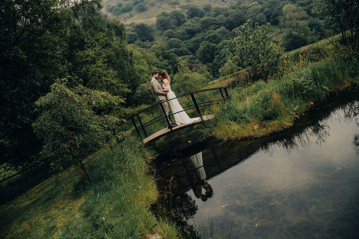 Wedding couple standing on rustic wooden bridge over pond with green hills and reflection in the water.