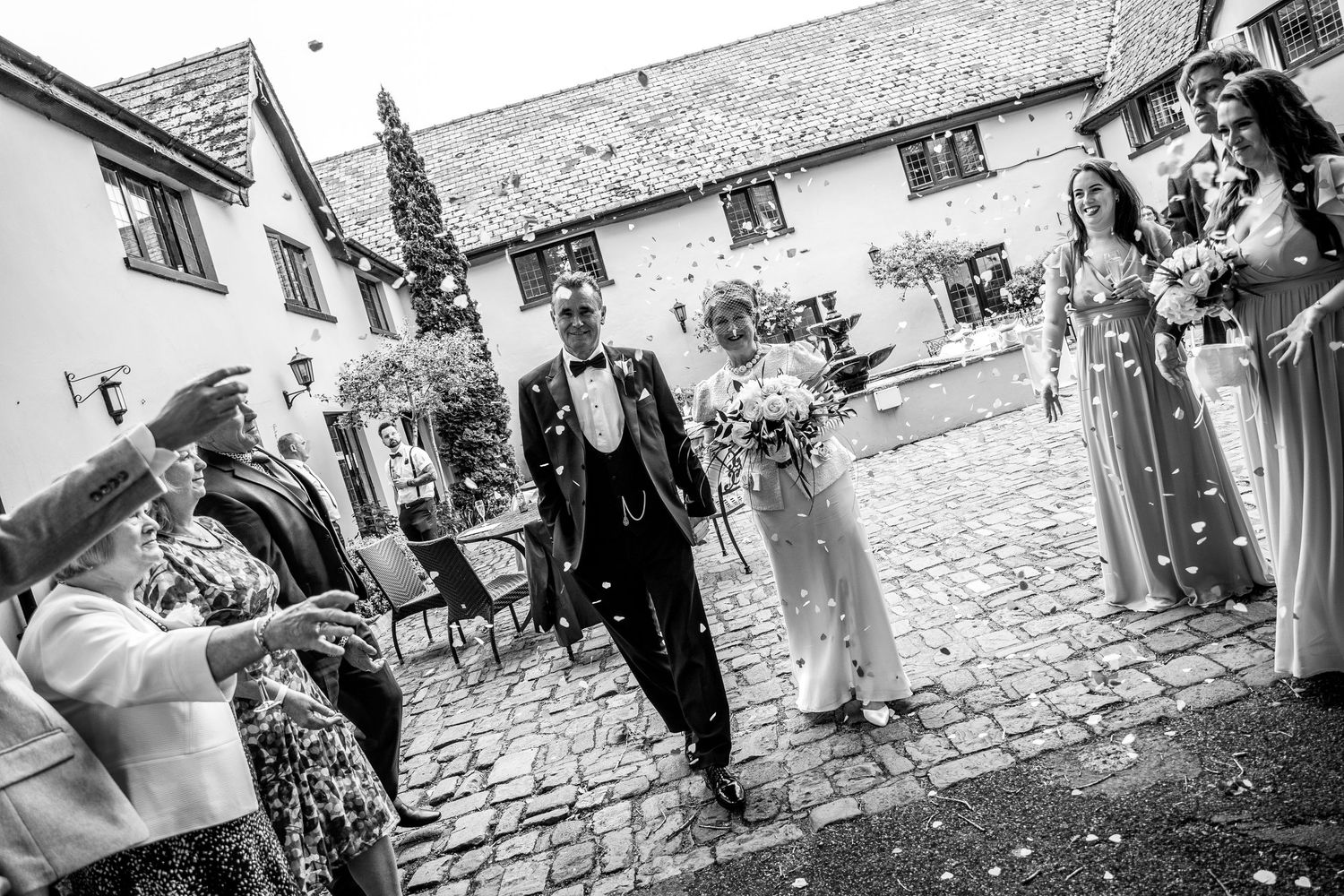 Wedding party celebrating with confetti throw in cobblestone courtyard.