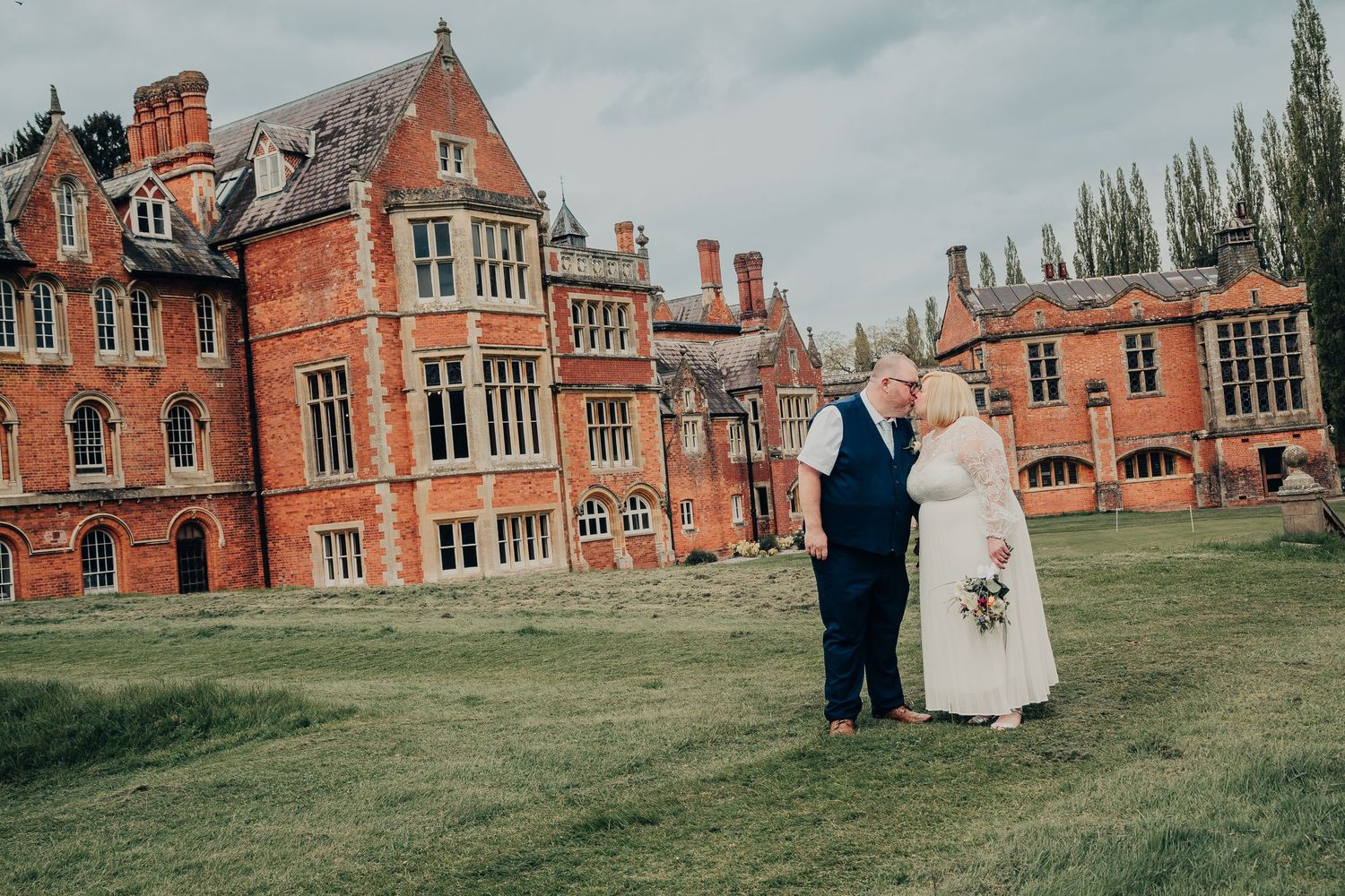 Couple stands together on the lawn of a grand Victorian red brick mansion with ornate architecture and tall trees.