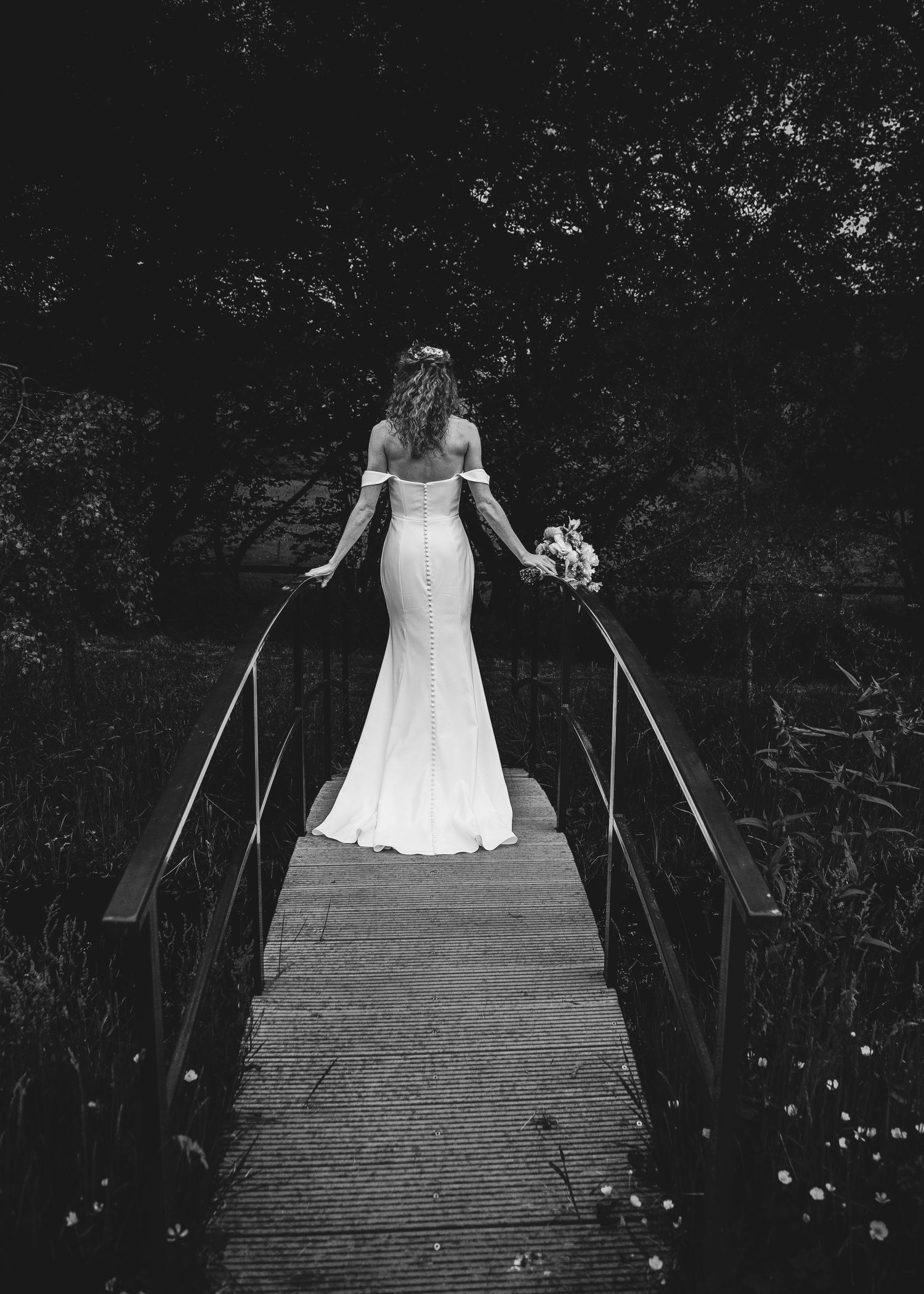 Bride in elegant fitted lace wedding gown standing alone on wooden footbridge in black and white photography.