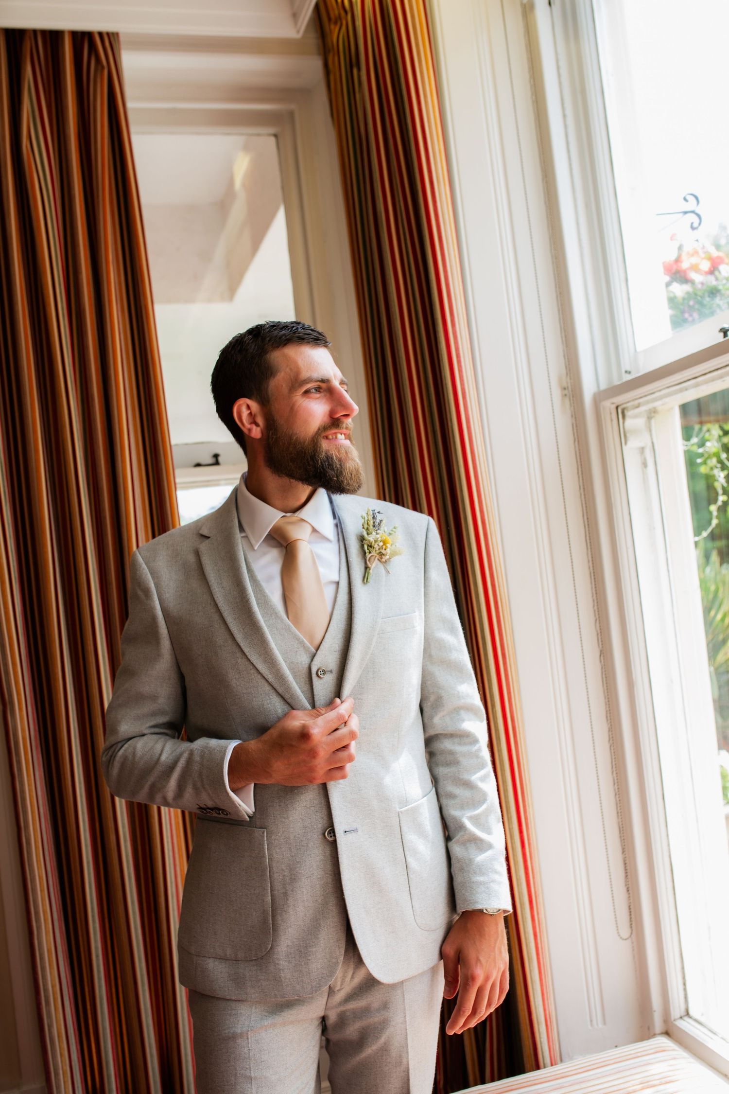 Groom in light grey suit poses thoughtfully by window.