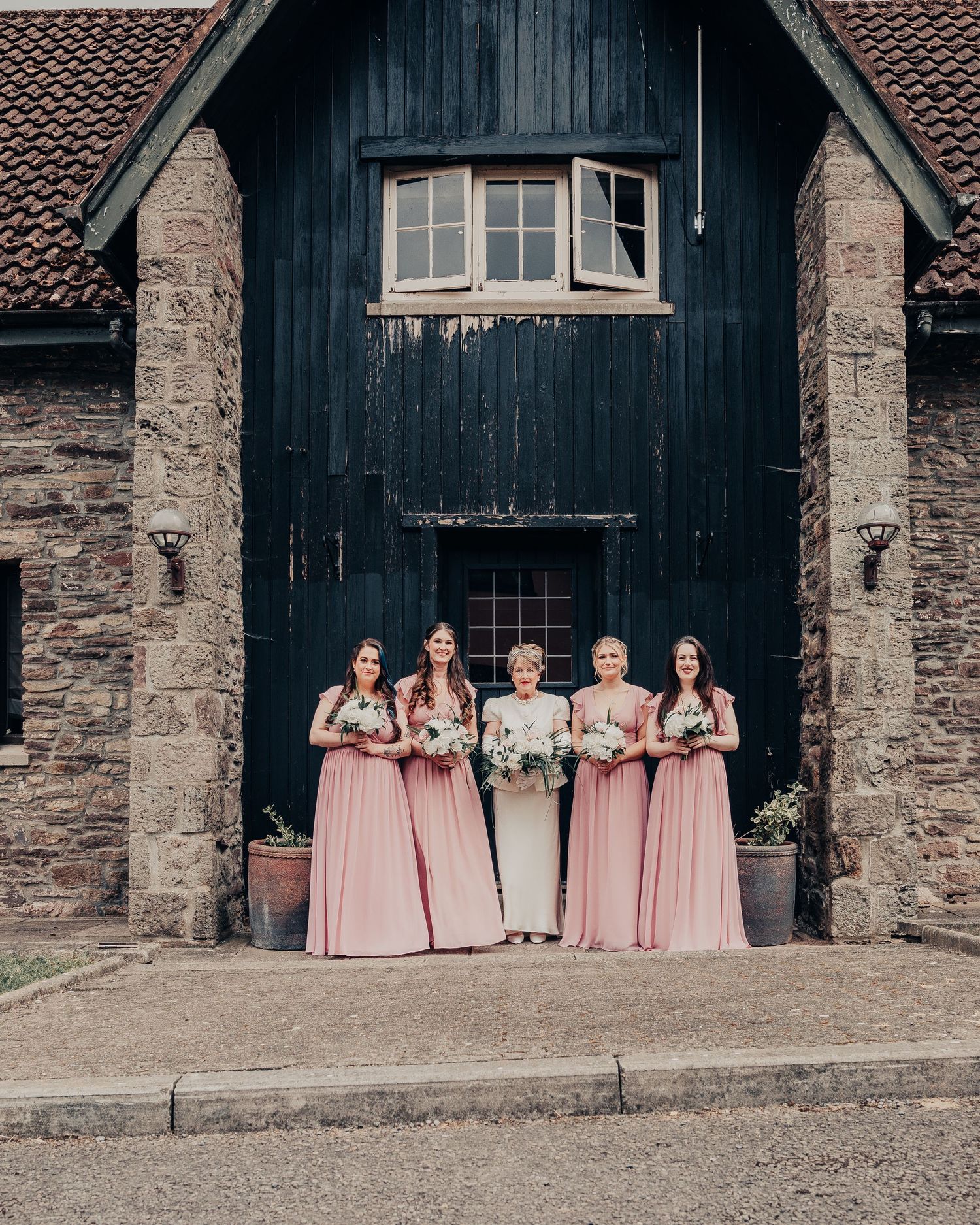 Wedding party stands in front of rustic stone and wood building wearing pink floor-length gowns.