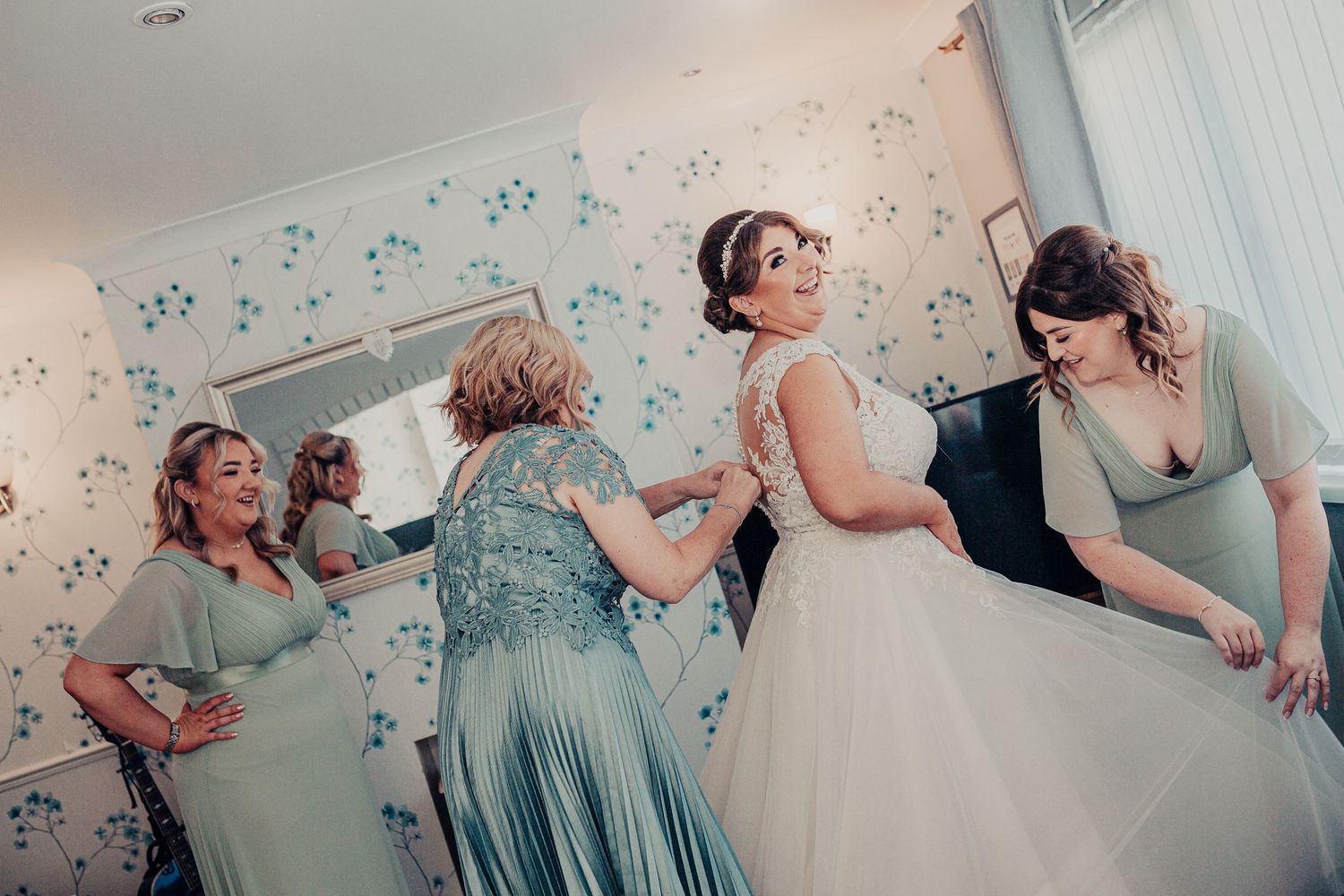 Getting ready scene with bridesmaids helping adjust wedding gown in floral wallpapered room.