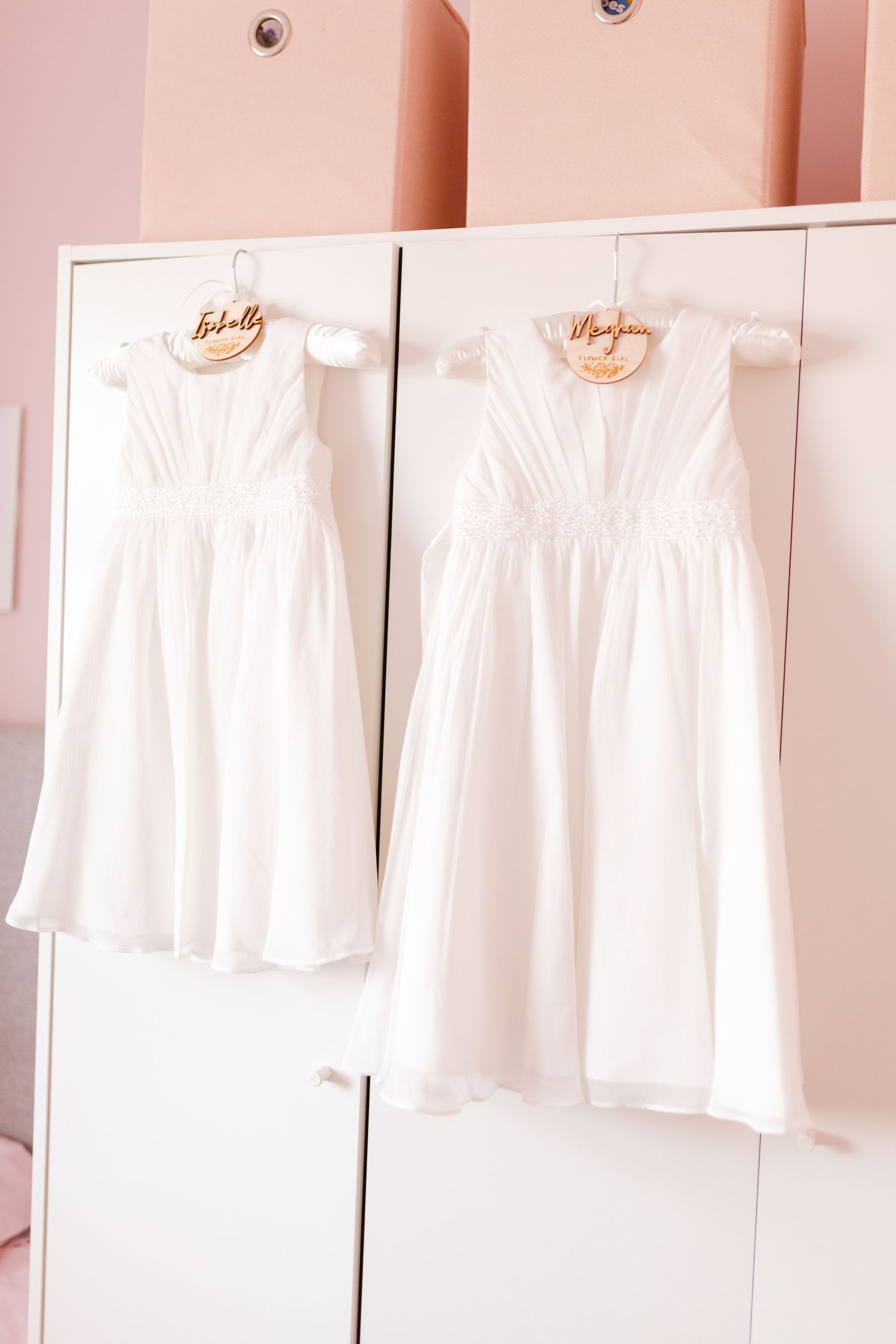 Two white flower girl dresses hanging on wooden hangers against a pink wall.