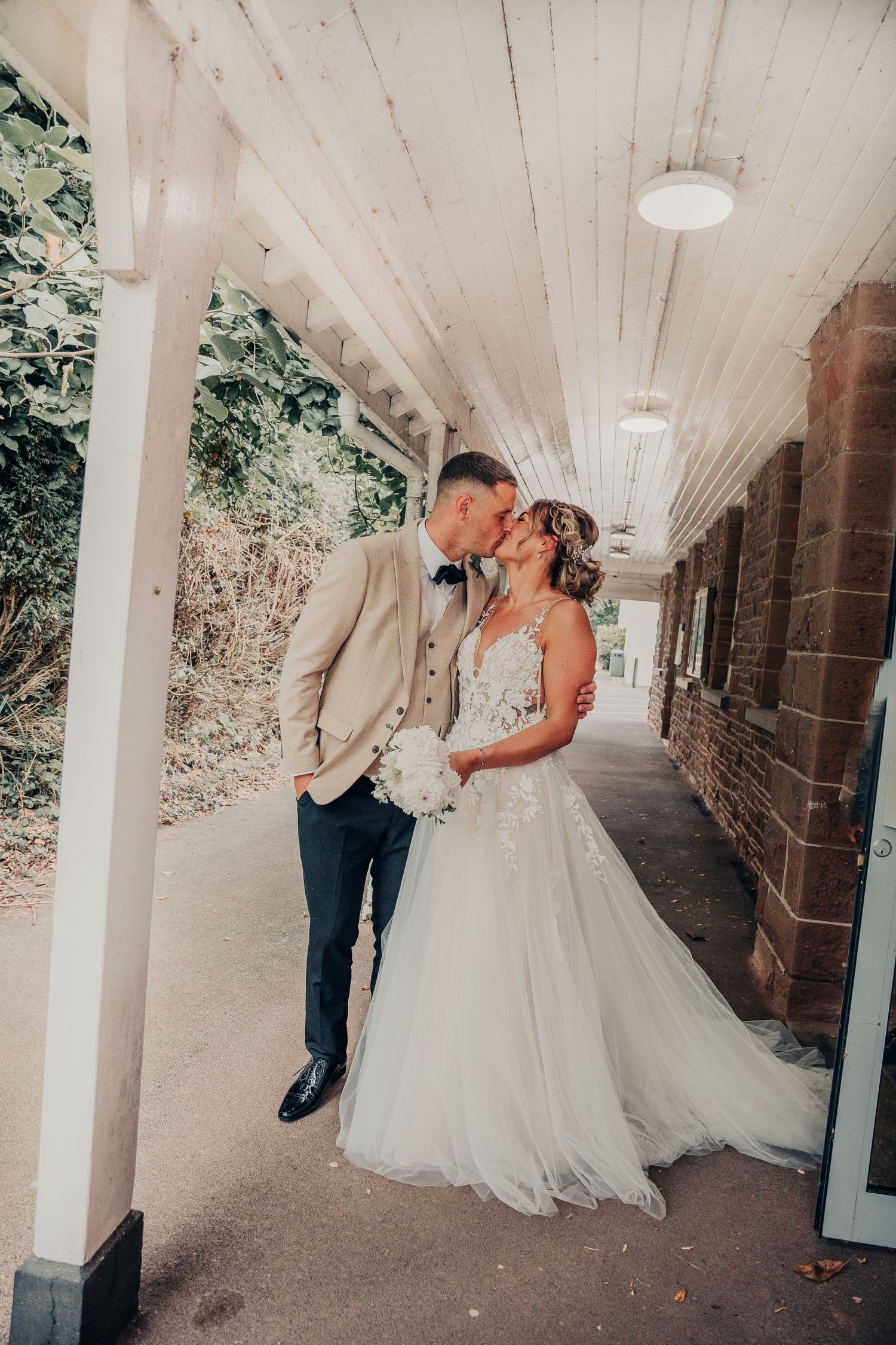 A romantic moment between a couple sharing a tender kiss in a covered outdoor walkway with white columns.