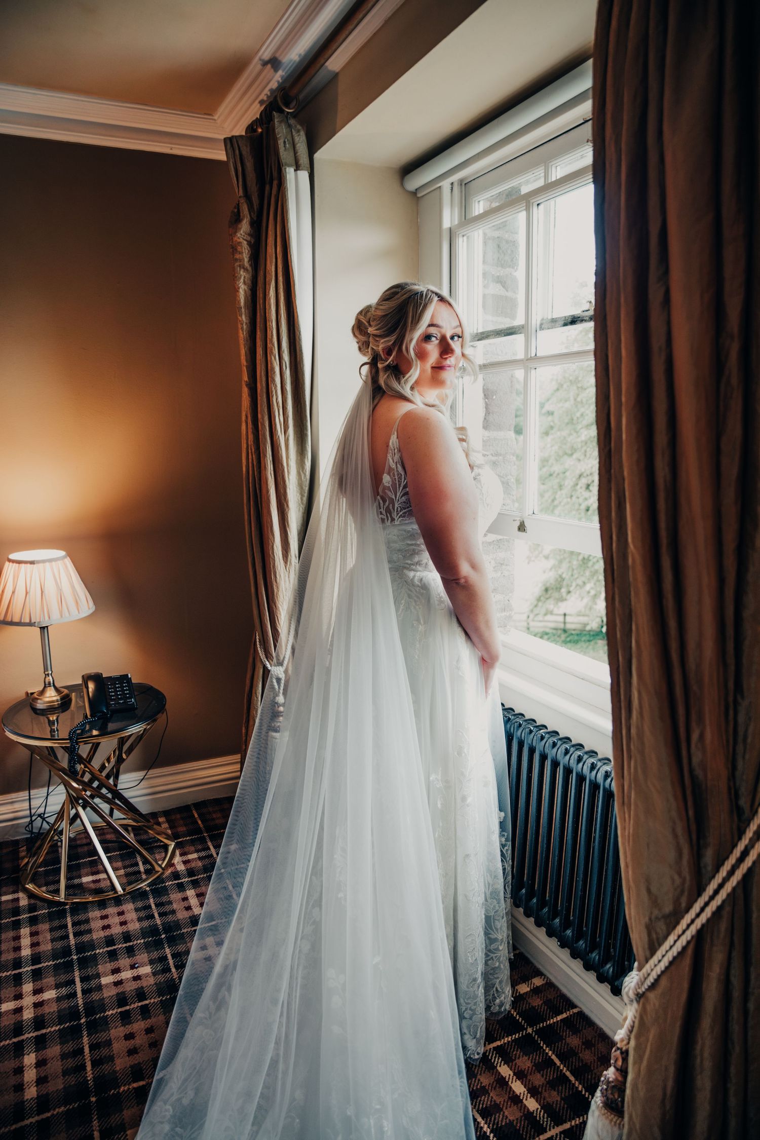 Elegant bride in flowing veil gazes out window while standing in traditional room with plaid carpet.