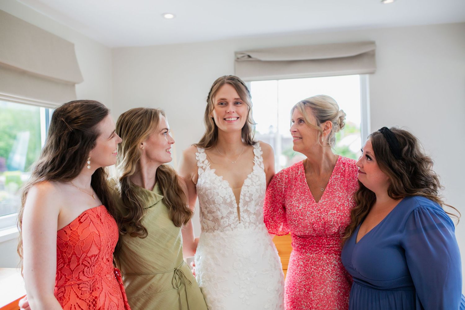 A group of women in colorful dresses gather around a bride in a white lace gown for a celebratory moment.