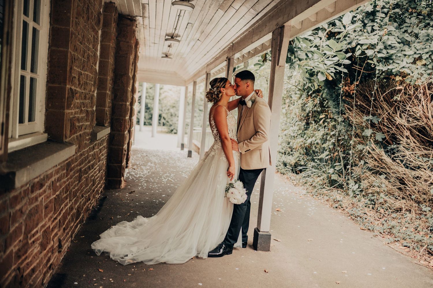 Two people in elegant wedding attire embrace in a sun-dappled outdoor garden setting with brick and white walls.