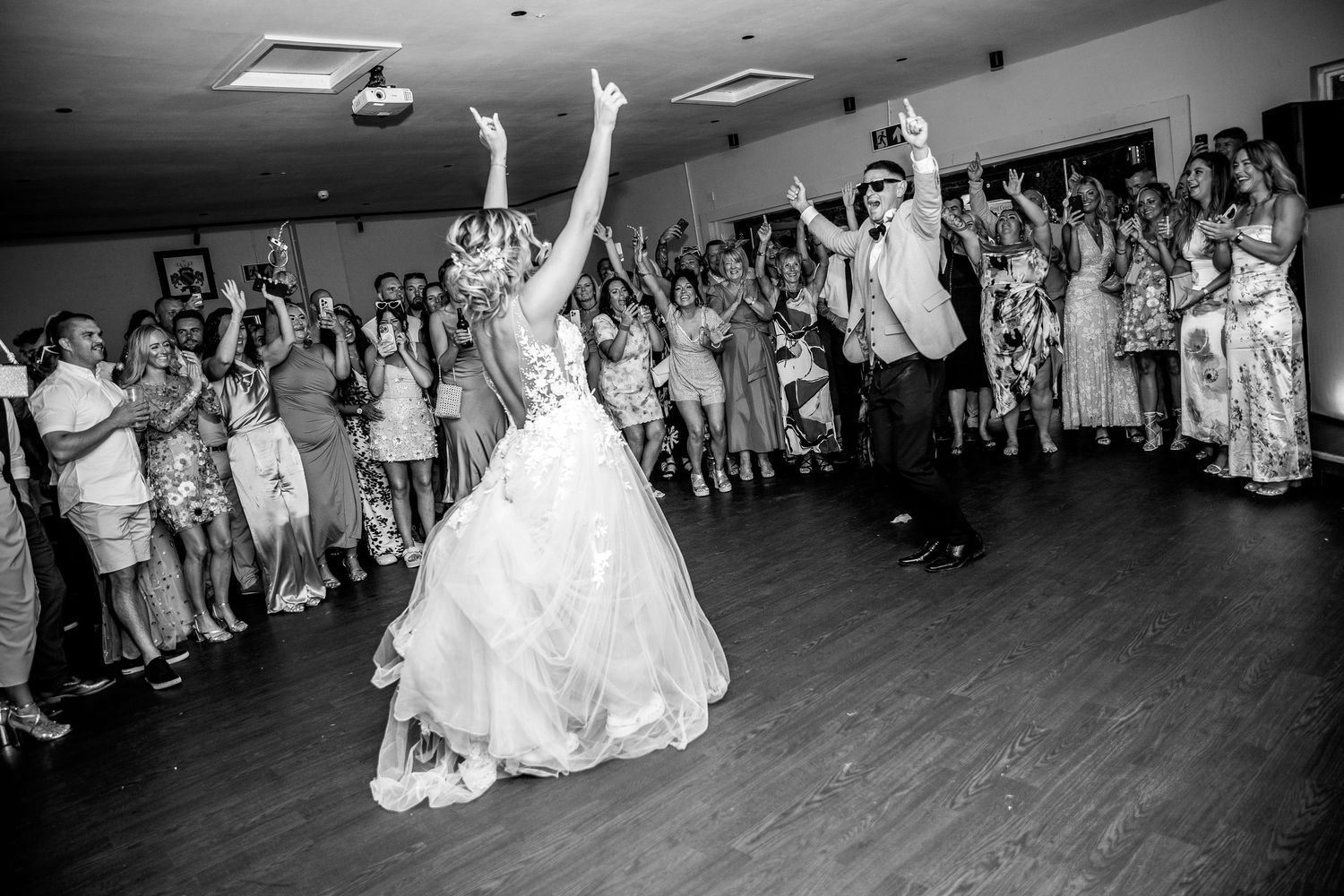 A newlywed couple dances energetically on a wooden dance floor surrounded by cheering wedding guests at a reception.