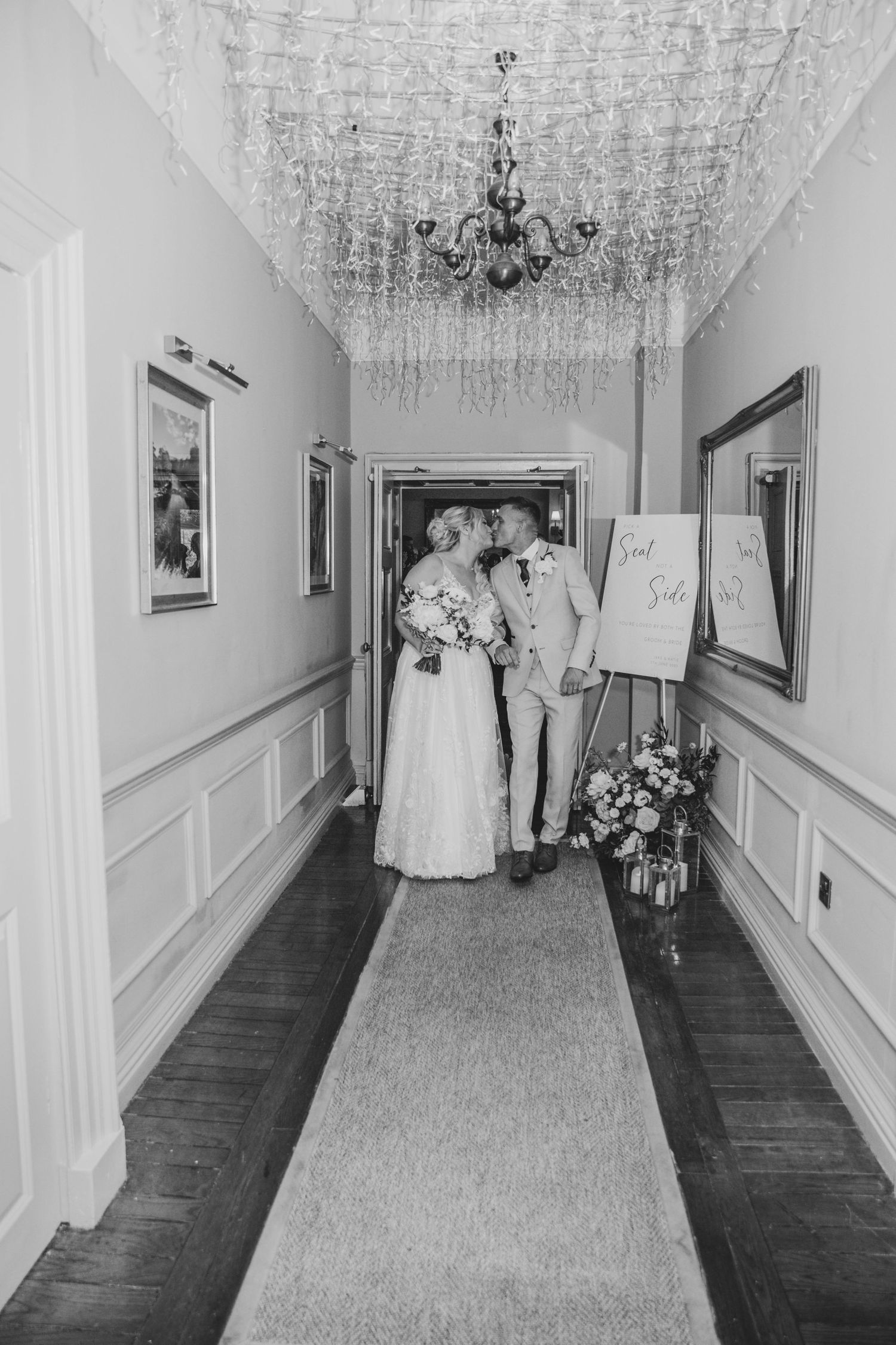 Black and white photo of newlyweds walking down an elegant hallway with chandeliers.