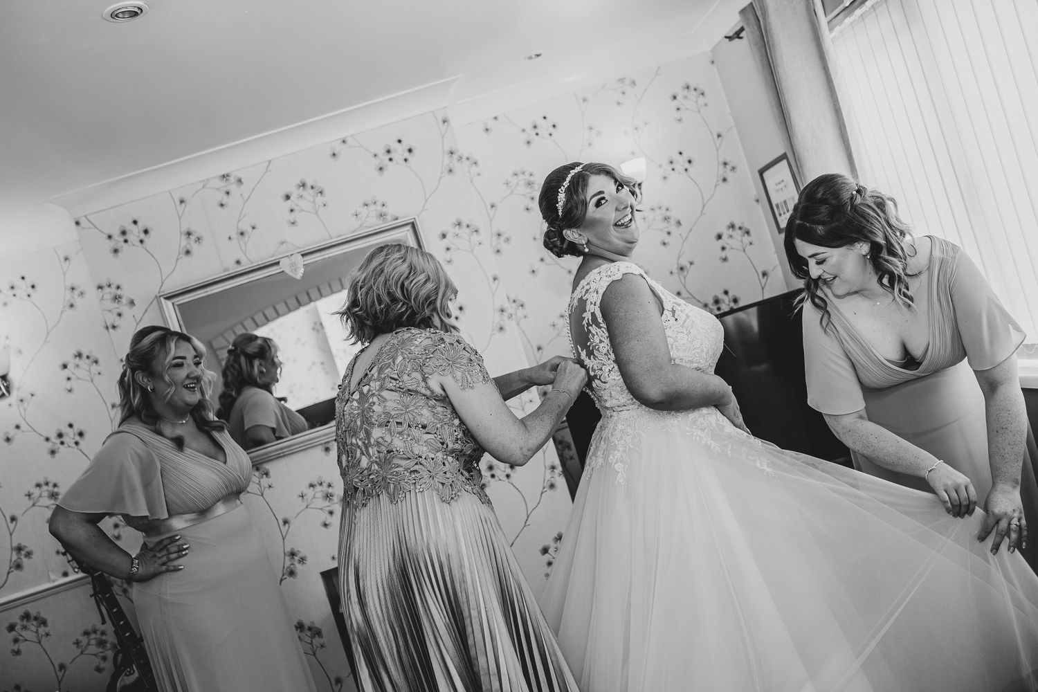 Black and white candid photo of wedding dress preparations with helpers in room.