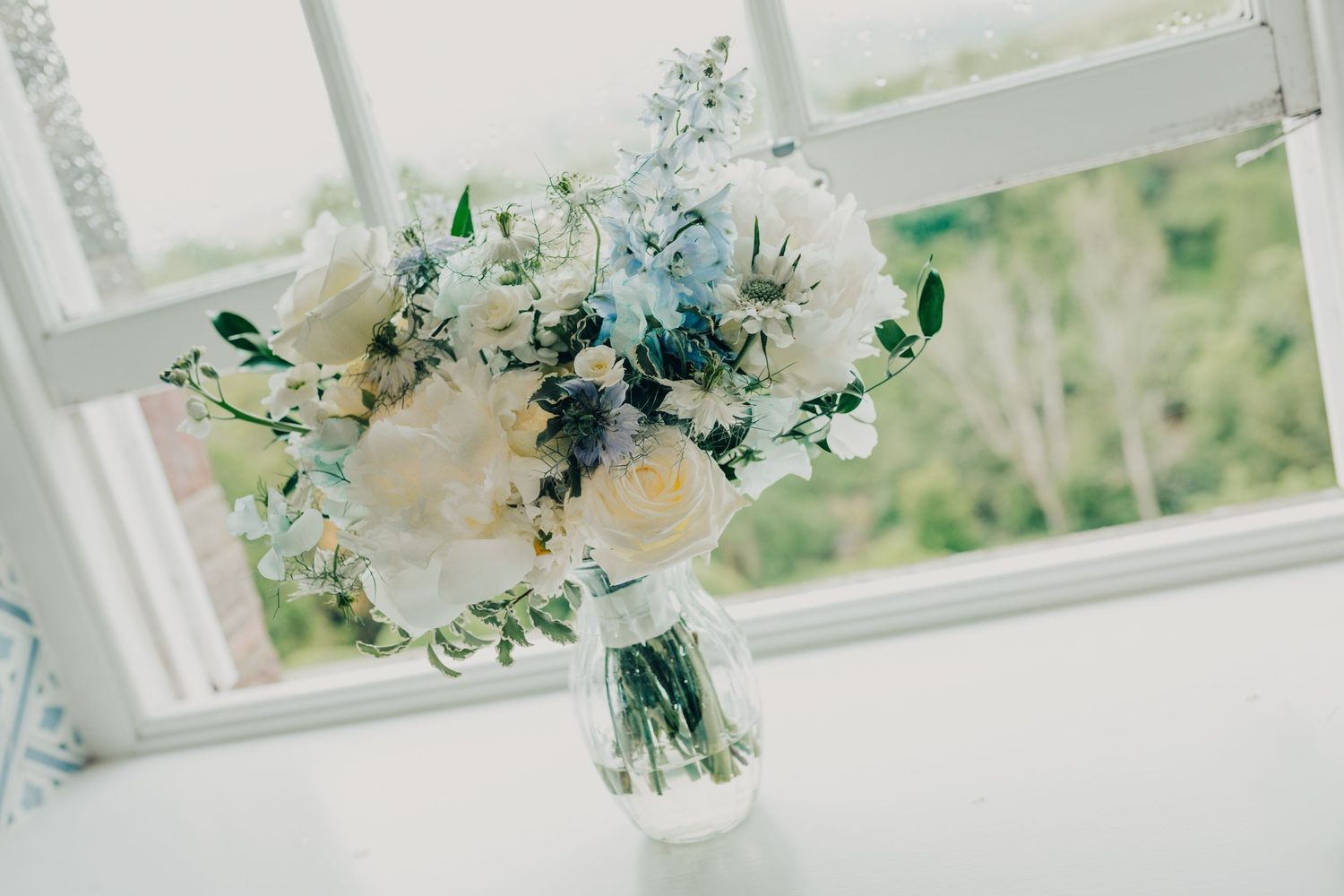 Delicate white and blue floral wedding bouquet in glass vase by window.