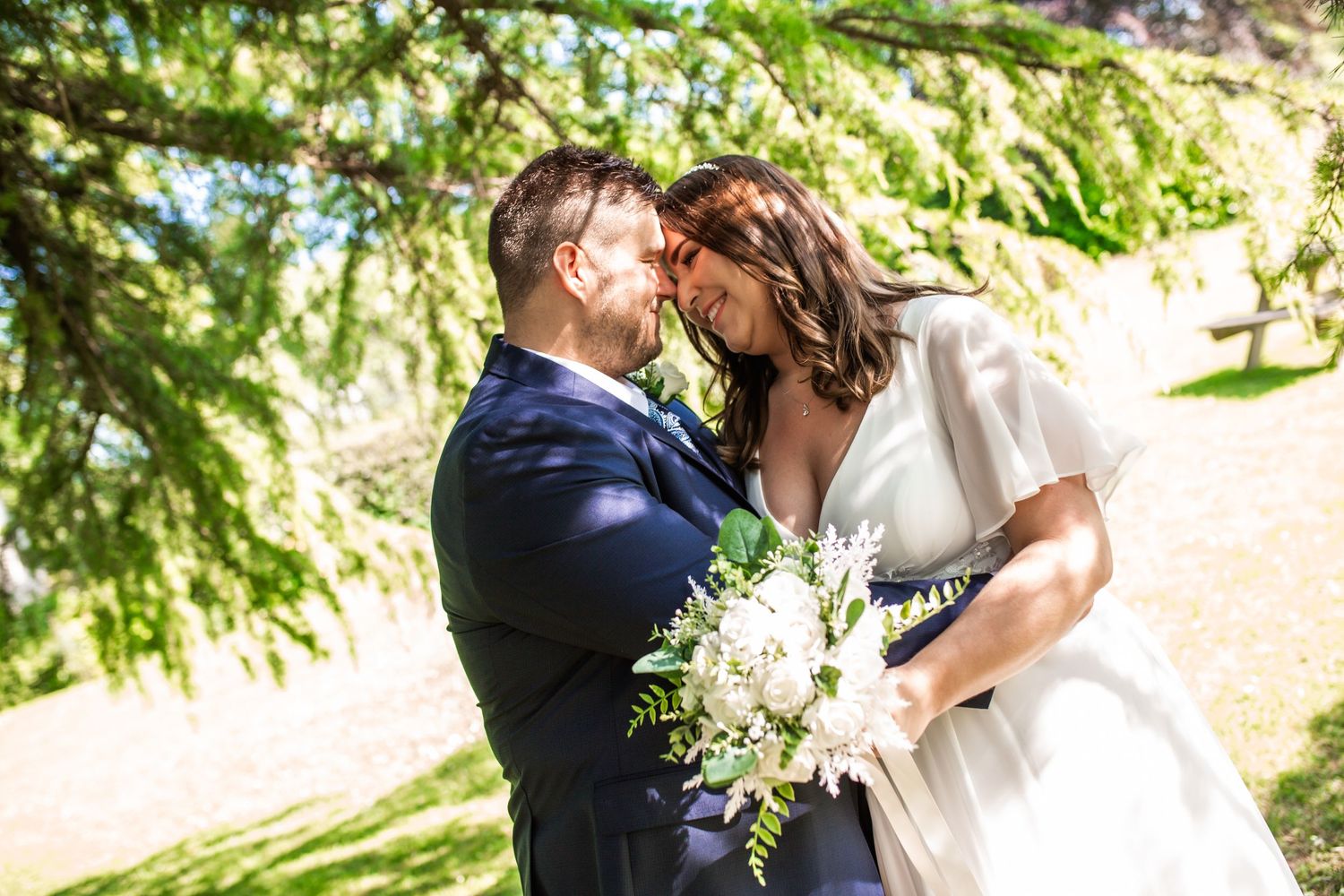 Romantic outdoor wedding portrait showing a tender moment between newlyweds with white floral bouquet.