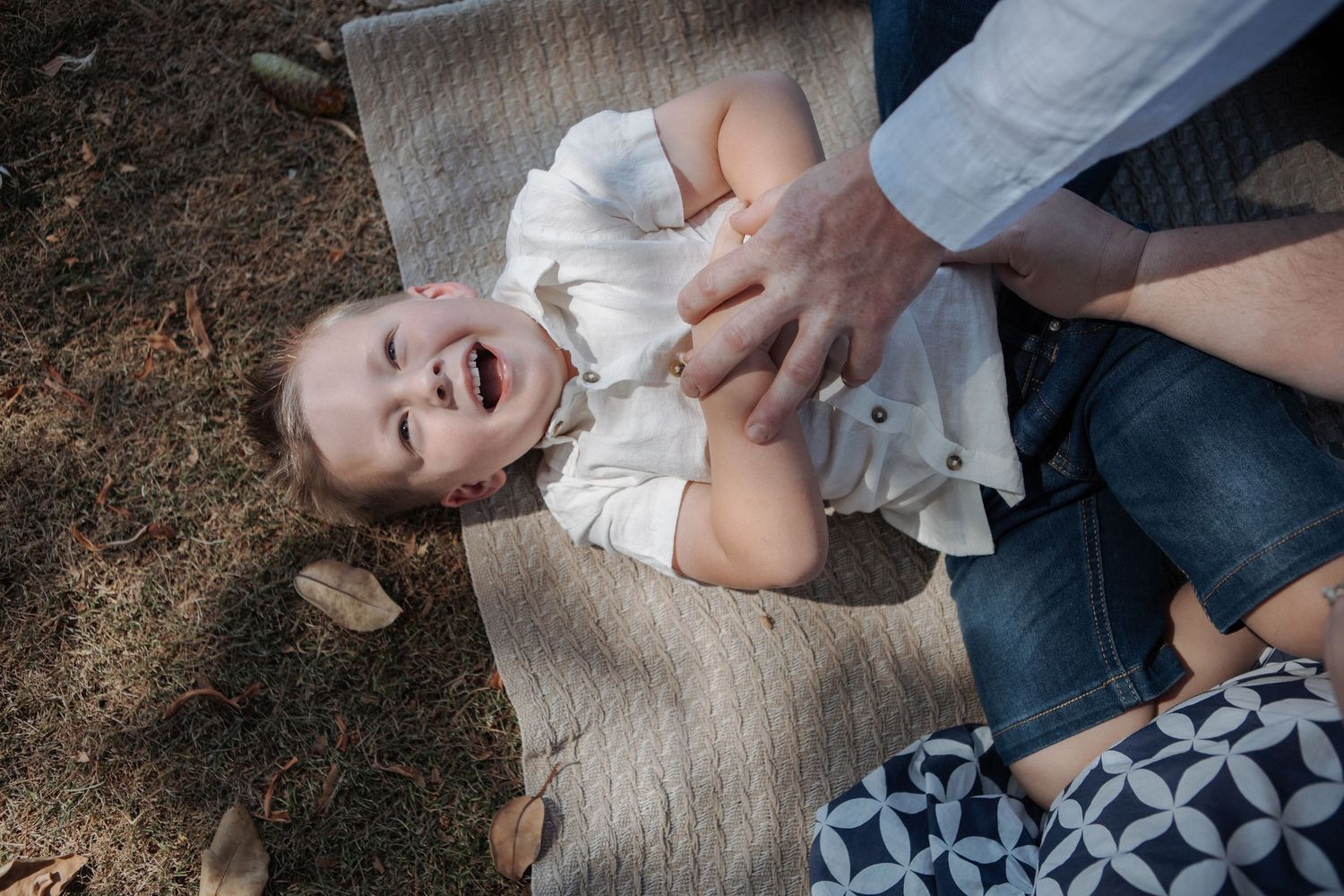 Joyful toddler in white shirt laughing while being tickled by parent on outdoor blanket.