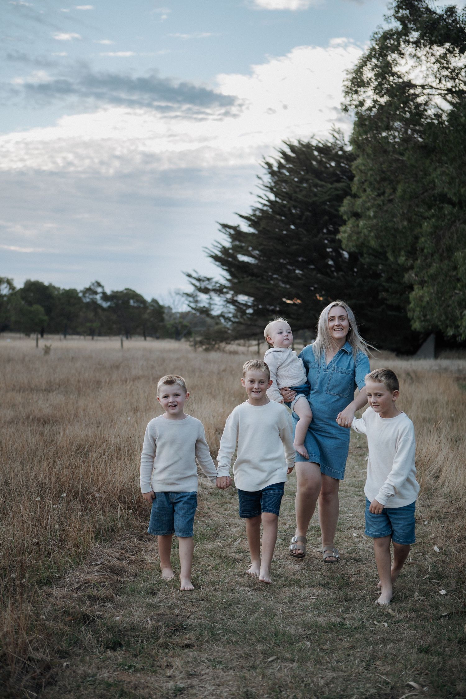 Mother in blue dress walks barefoot through golden field holding baby while three boys walk ahead.