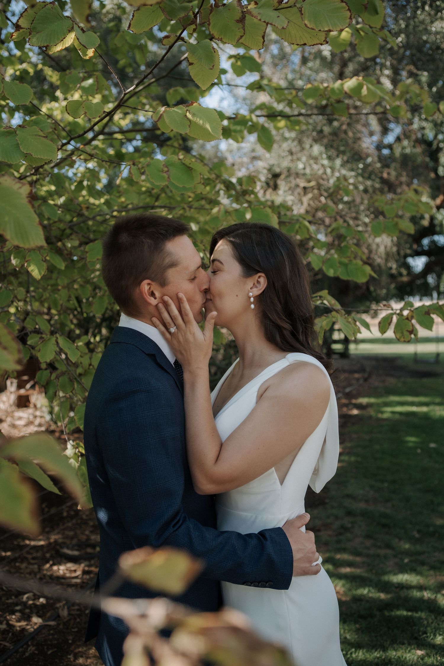 A bride and groom share a romantic kiss under lush green trees in an outdoor garden wedding setting.