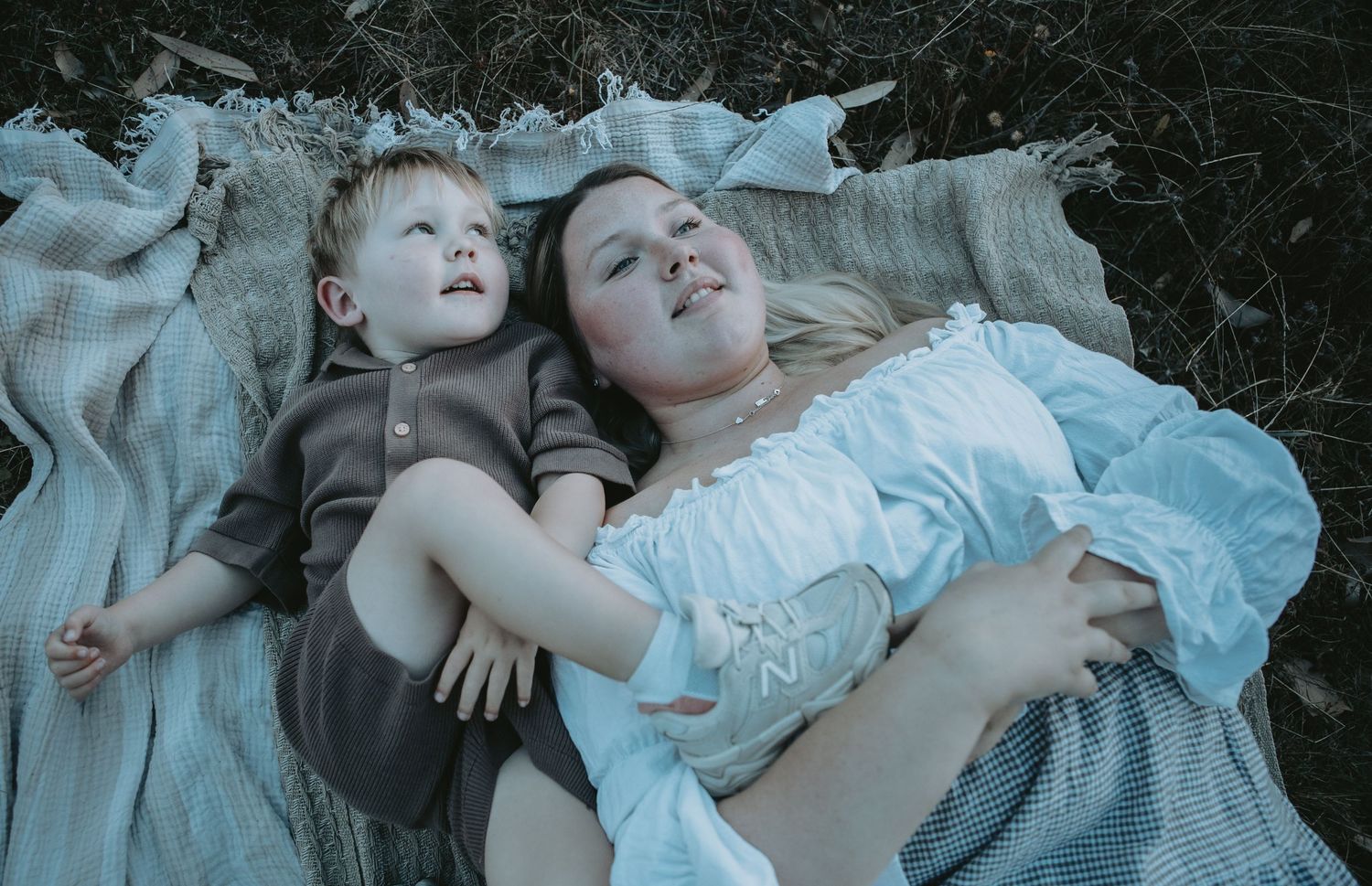 A mother and young toddler lie together on a blanket in a natural outdoor setting.