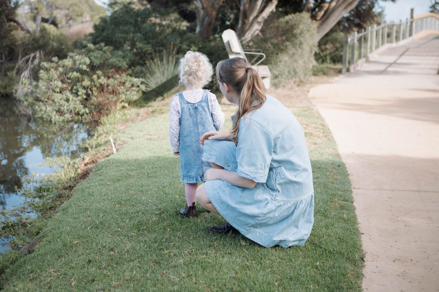 Pregnant woman in blue dress crouching beside toddler in denim overalls on green grass near a pond.