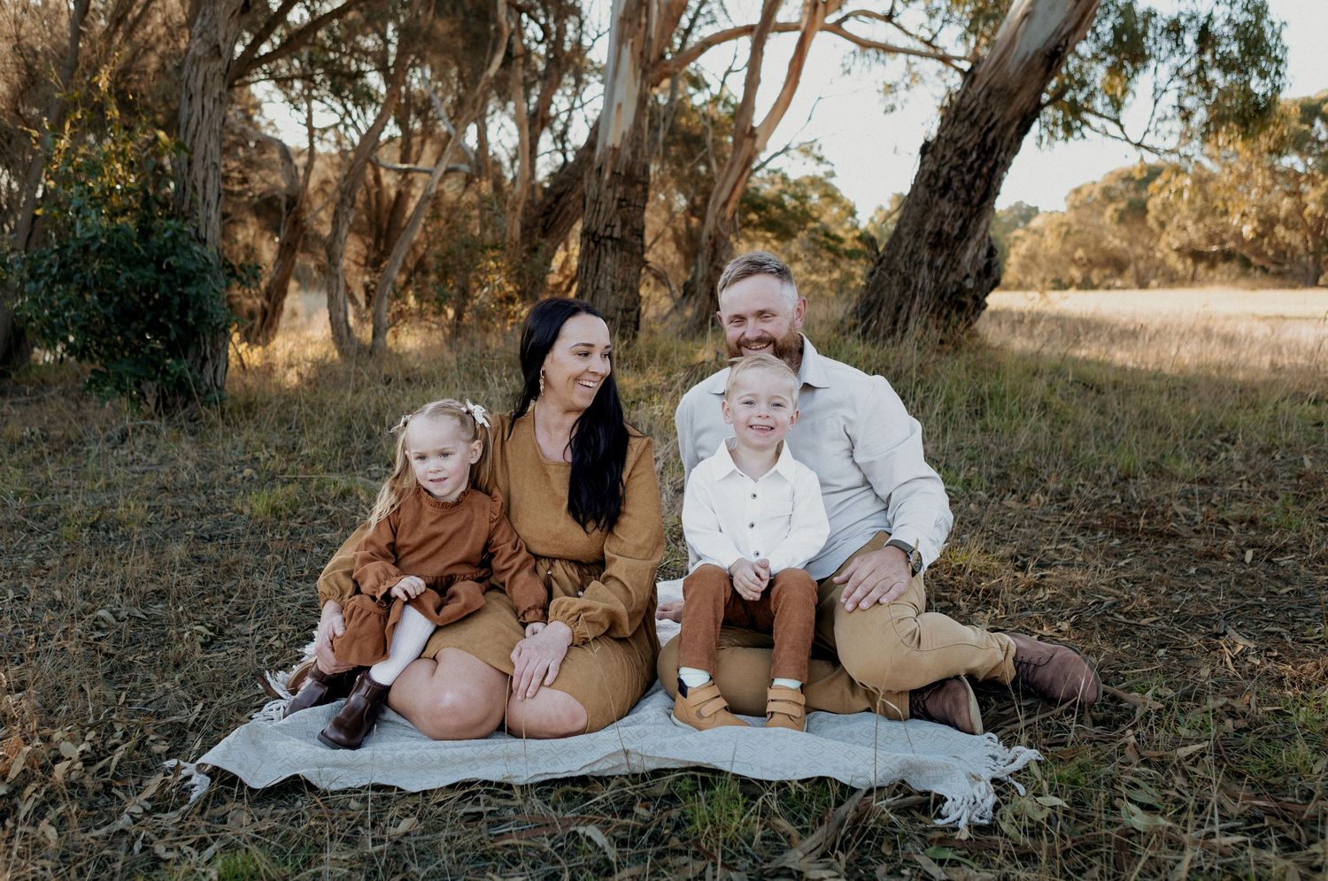 Family of four sitting on a blanket in a natural outdoor setting, wearing warm earth-toned outfits.