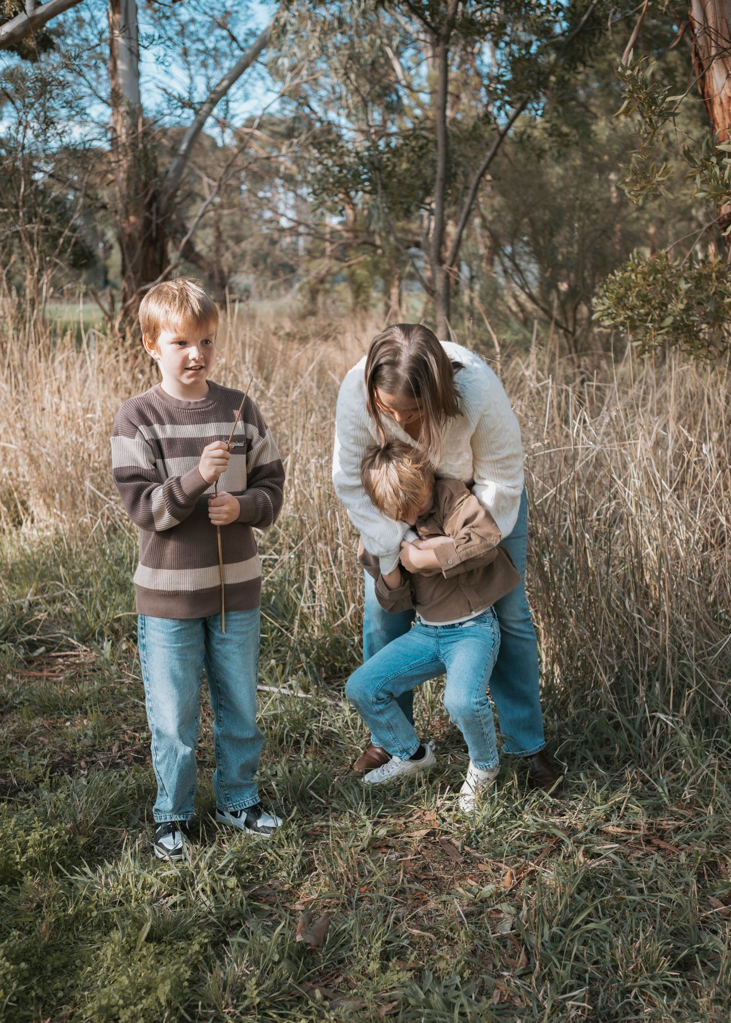 A mother hugs her toddler while an older boy holds sticks in a grassy woodland setting.