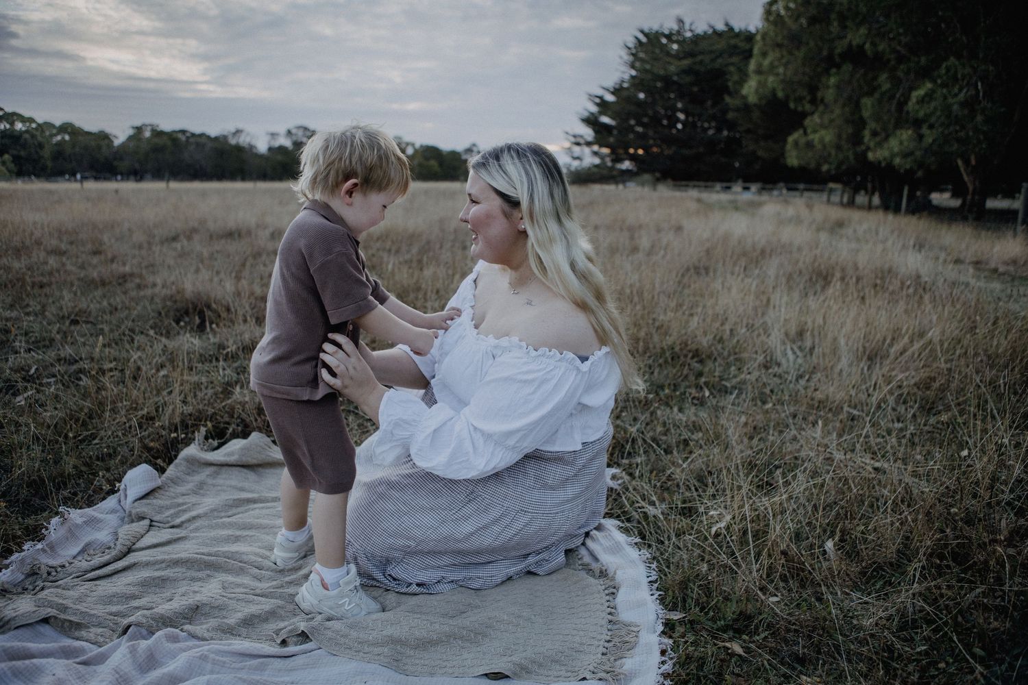 Mother in white off-shoulder top sits on blanket in golden field interacting warmly with her toddler son.