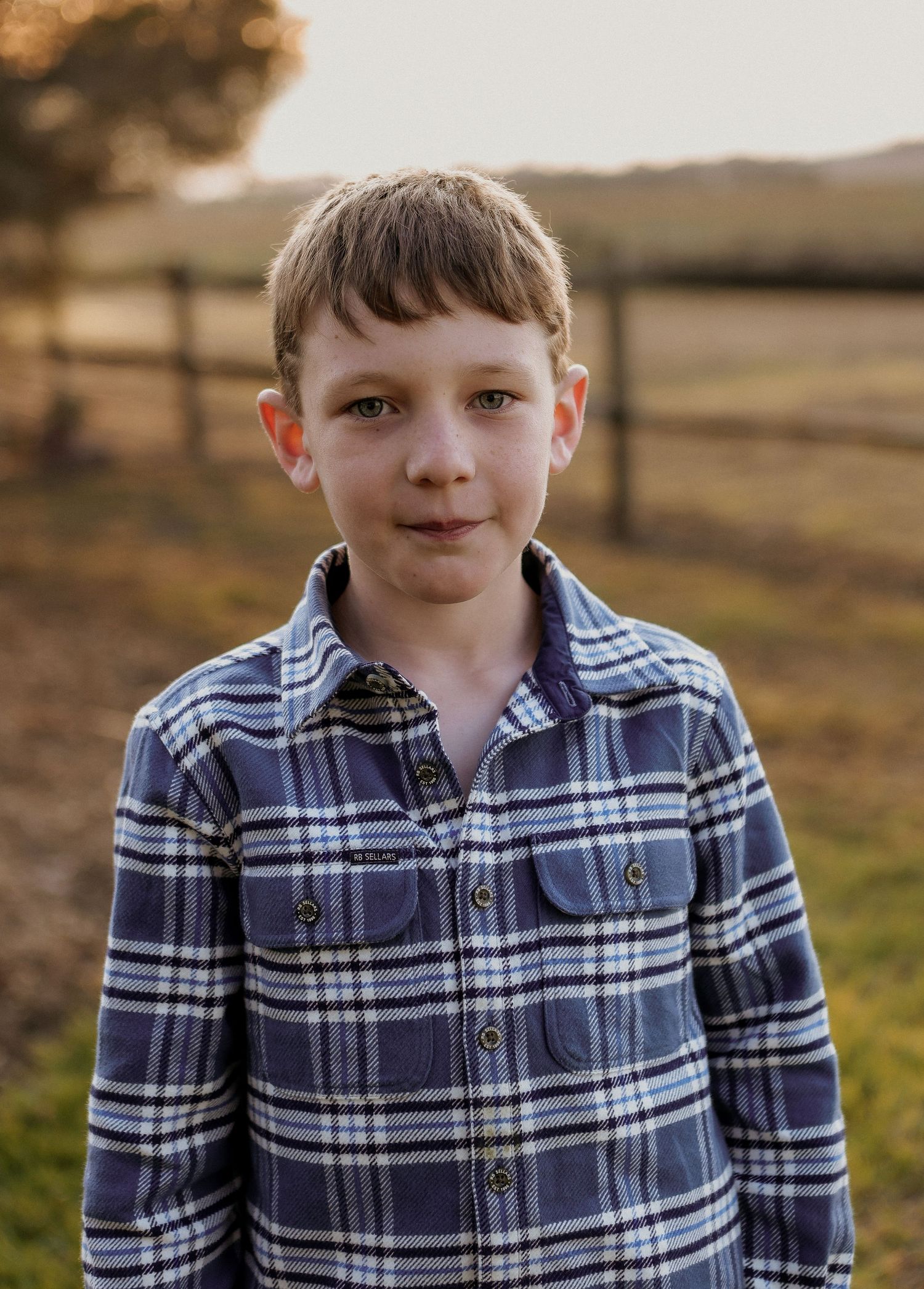 Young boy in blue plaid flannel shirt smiling outdoors at golden hour with rural fence background.