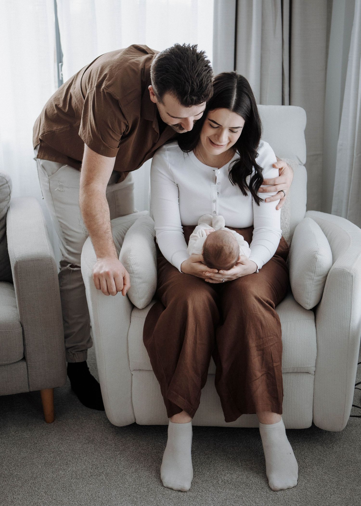 A couple smiles at their newborn baby, mother seated in a white chair, father leaning over her shoulder in a cozy home.