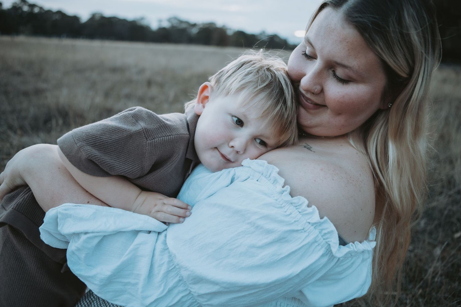 A mother tenderly embraces her young toddler son in a golden field at dusk.