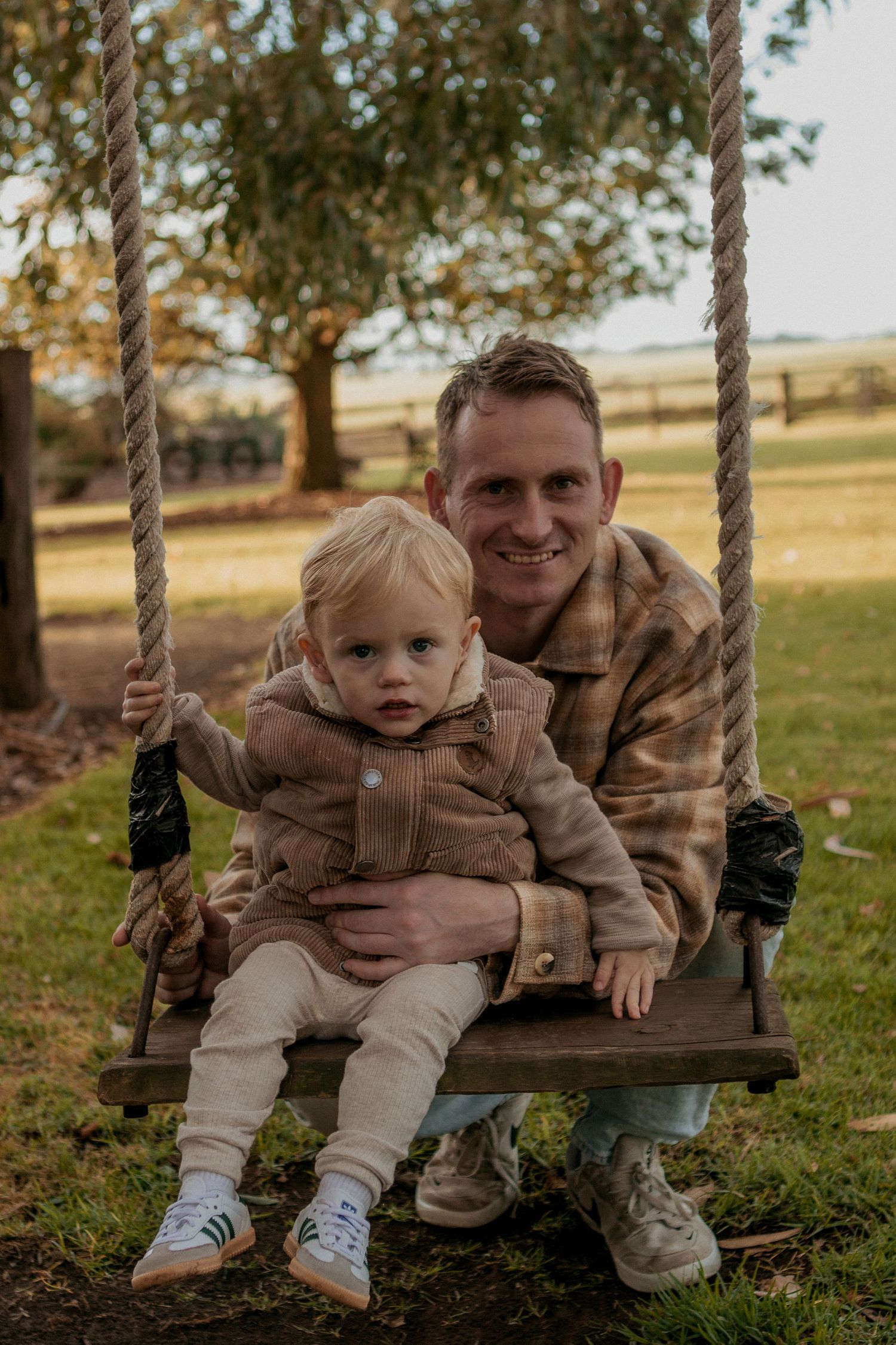 A smiling father sitting on a wooden rope swing with his young toddler in a rural setting.