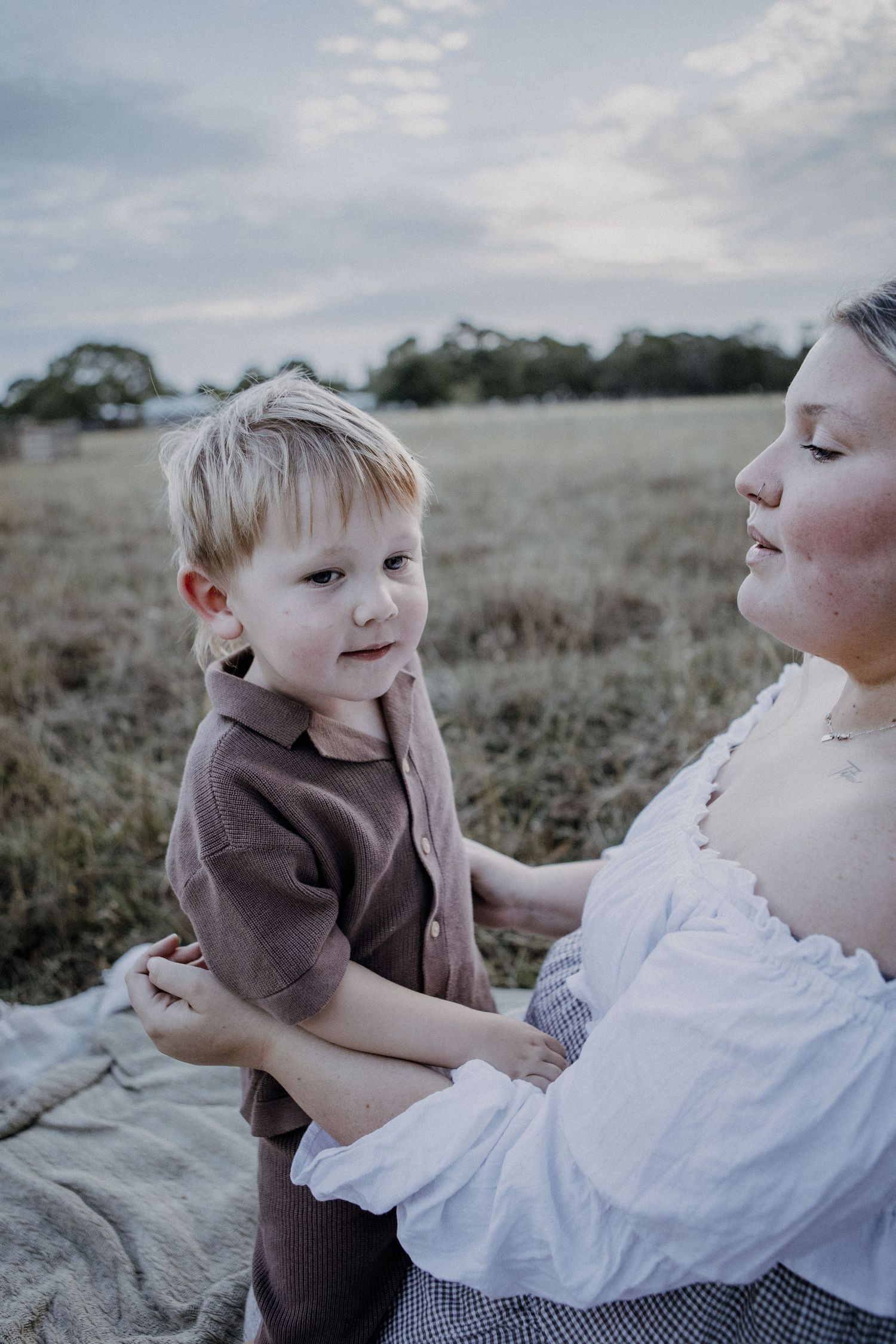 A young blonde toddler boy sits in his mothers arms on a blanket in a golden field.